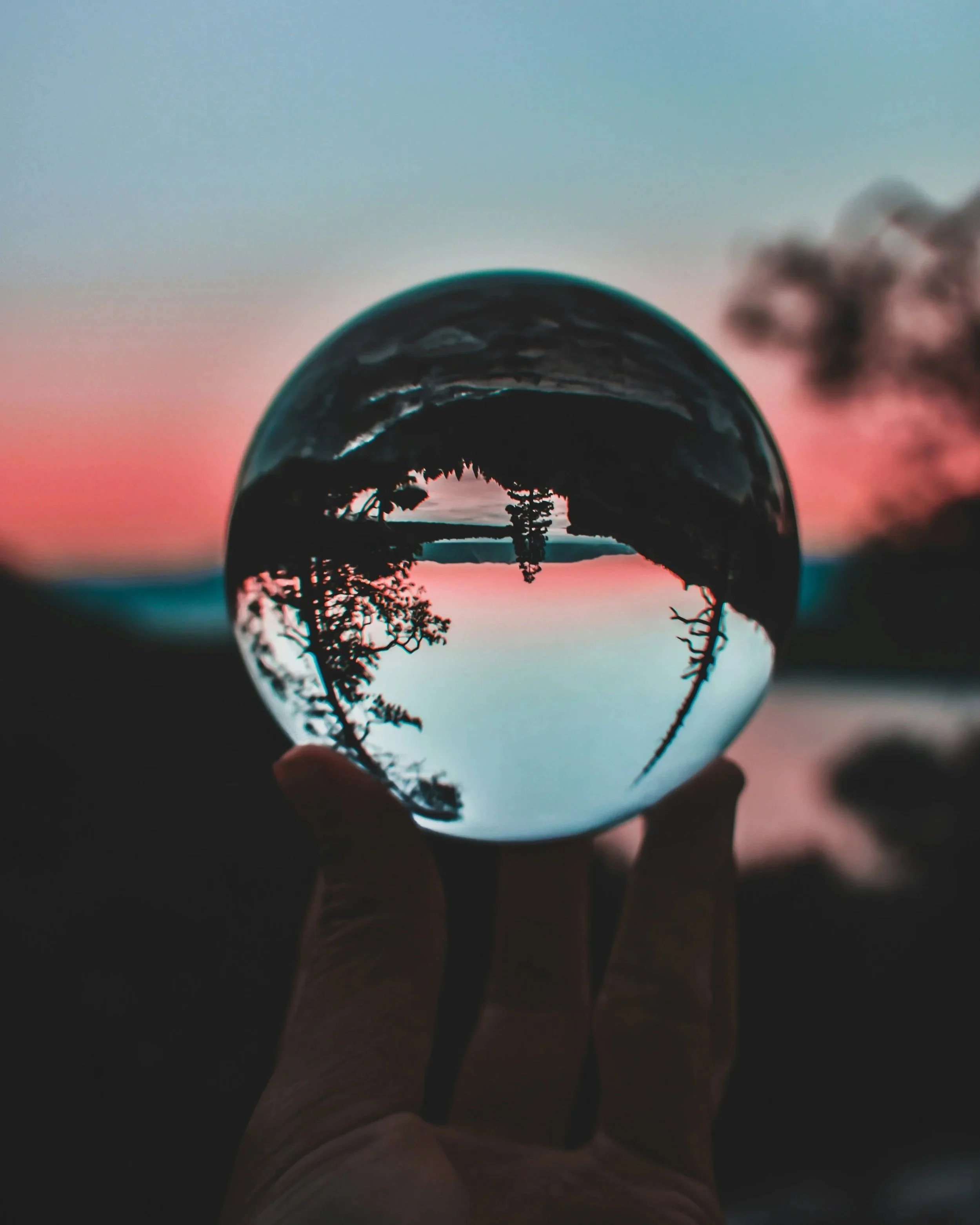 Hand holding a glass sphere reflecting a sunset over a lake with trees.