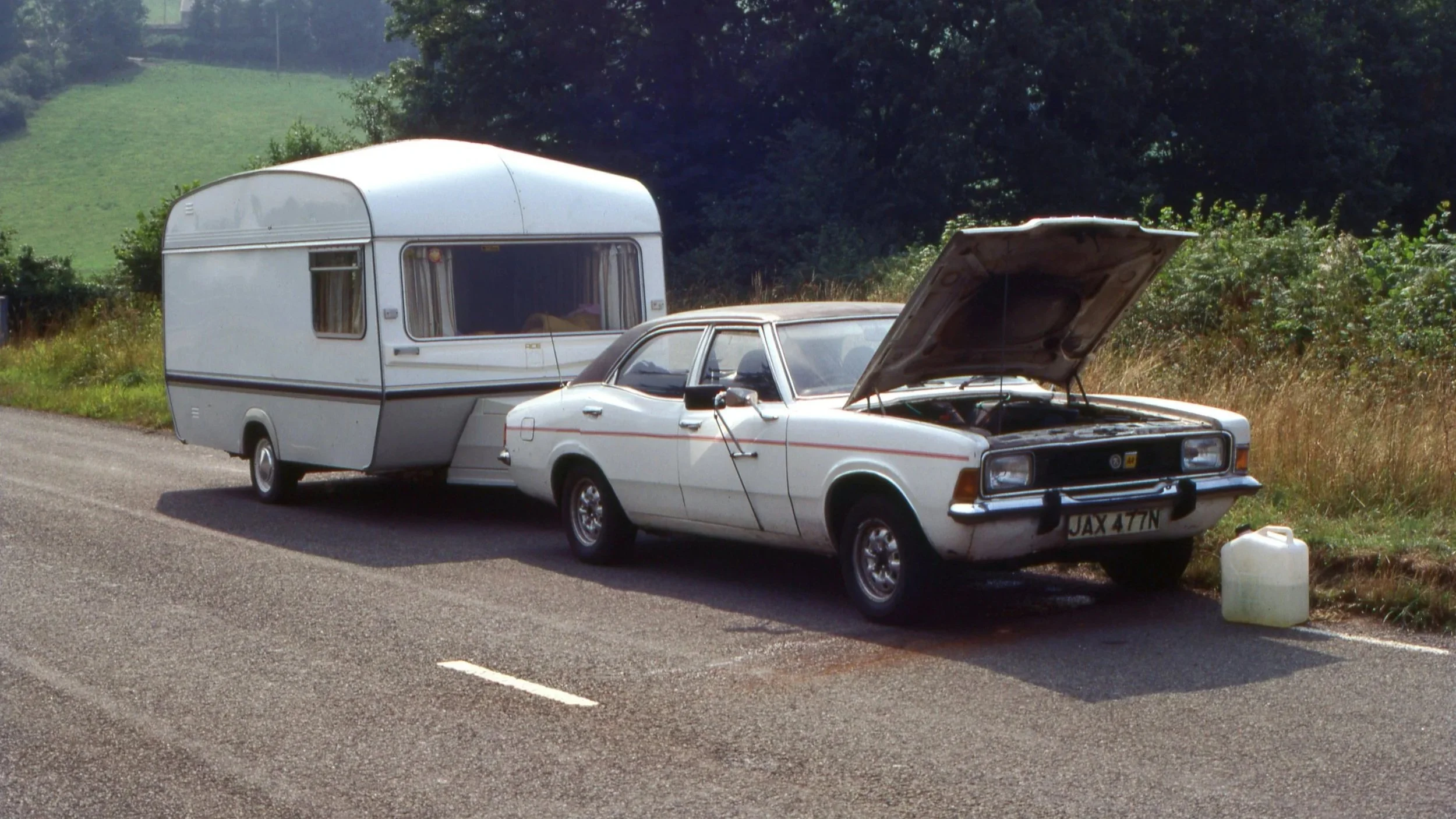 A white car with its hood open, towing a small white camper trailer on the side of a rural road during daytime.