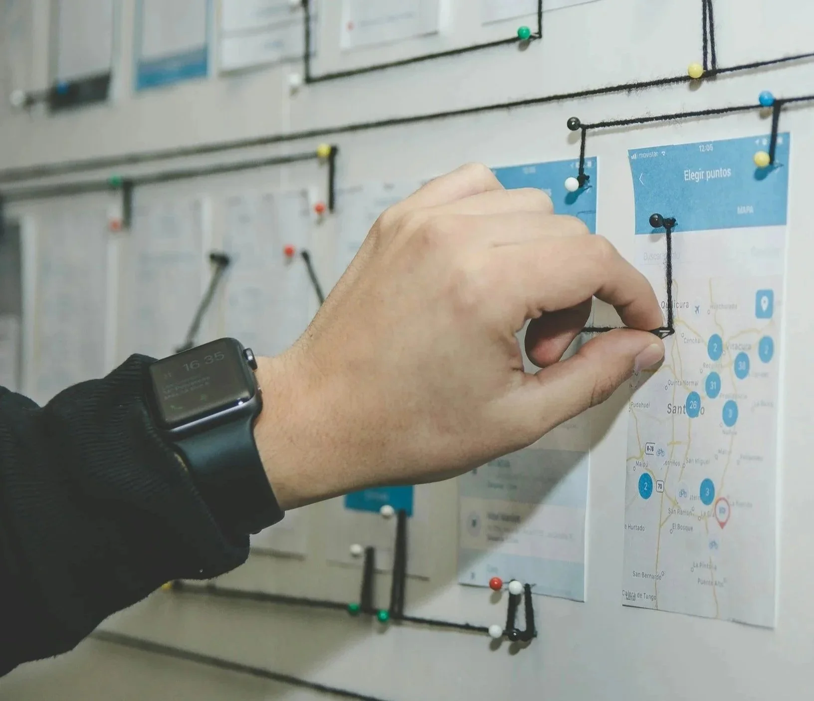 A person pinning a black string to a bulletin board that has a map and other papers pinned with colorful pushpins.
