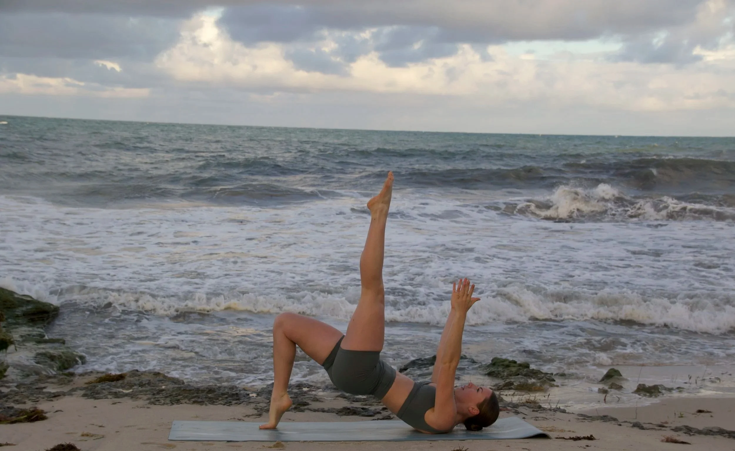 A woman practicing yoga on a beach, lying on her back with one leg extended vertically and her arms raised, facing towards the ocean with waves and a cloudy sky in the background.