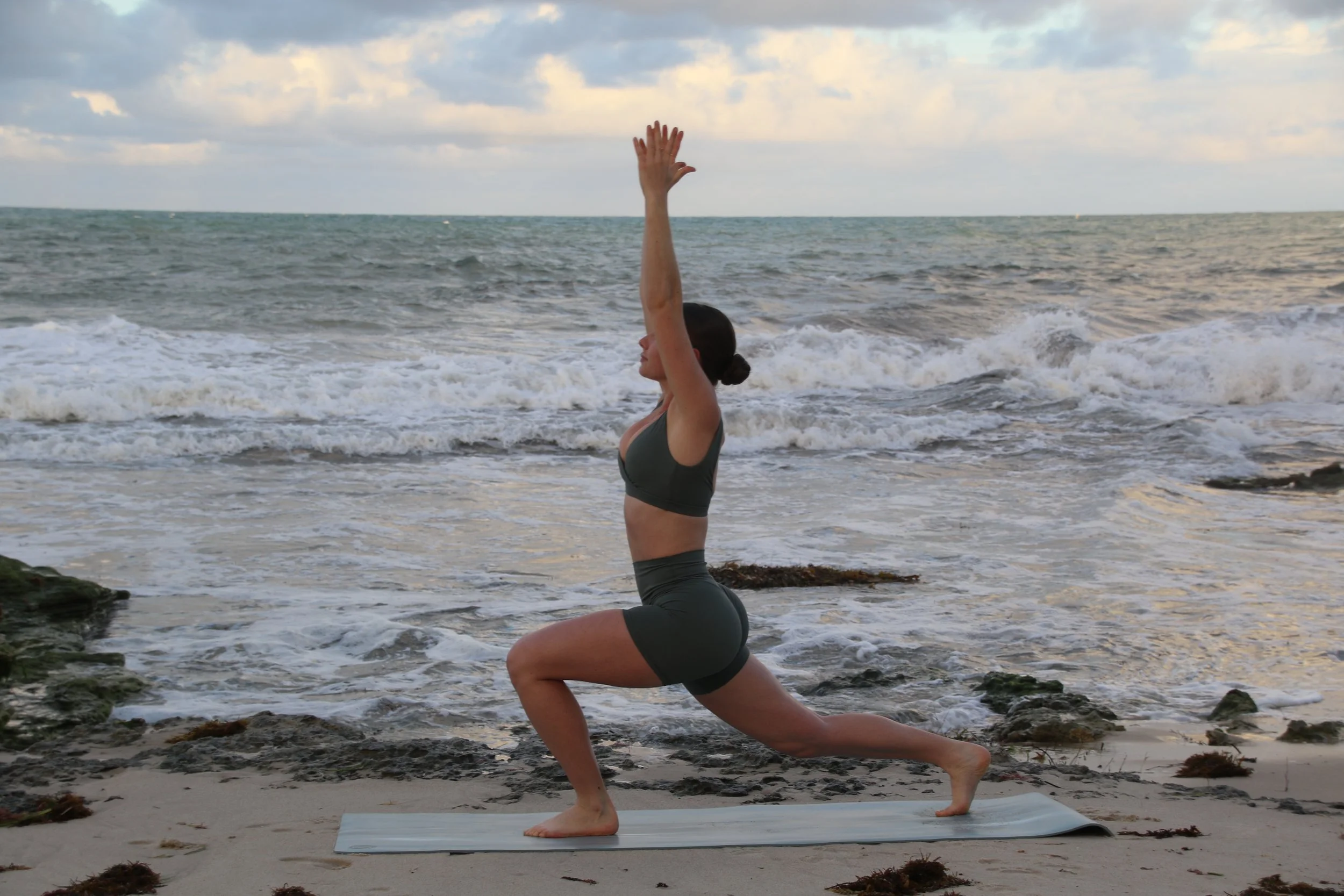 A woman practicing yoga on a beach near the ocean, performing the Warrior I pose with one knee bent, one leg extended back, and arms raised overhead.