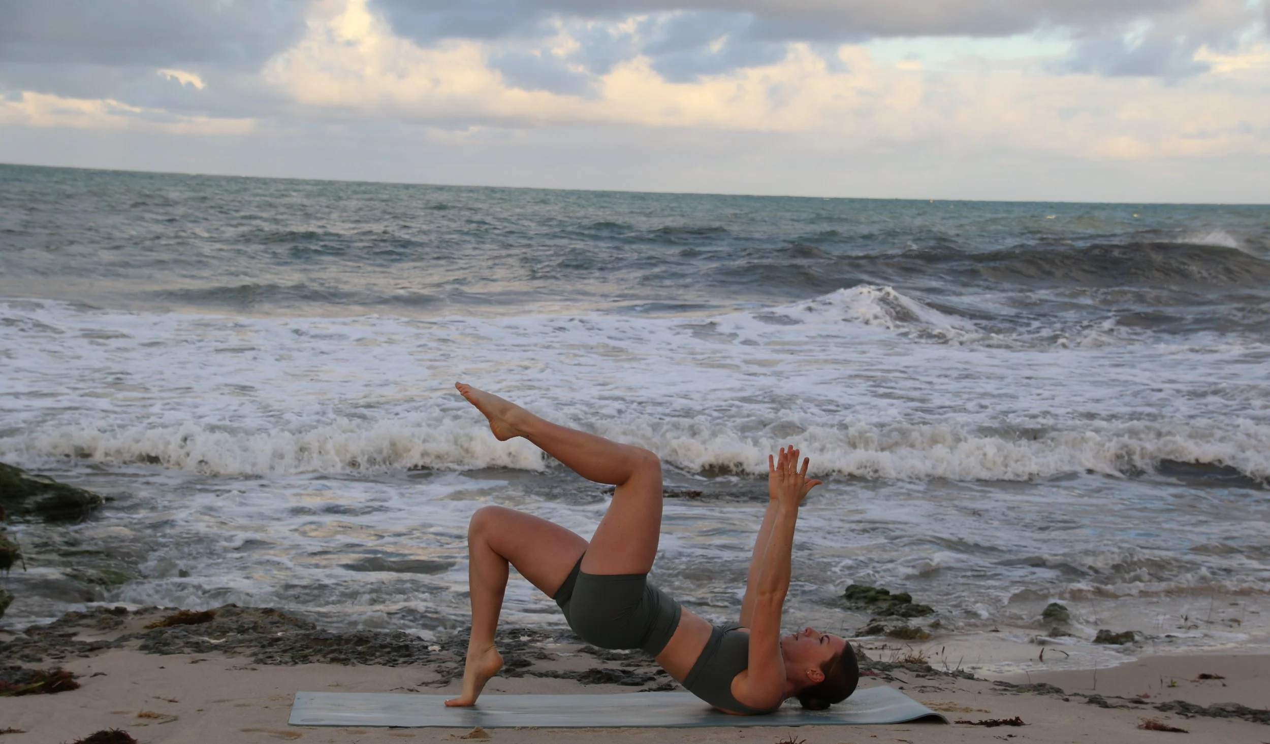 Woman practicing yoga on a beach mat on the sand, performing a pose with legs raised and one arm extended, ocean waves in the background under cloudy sky.