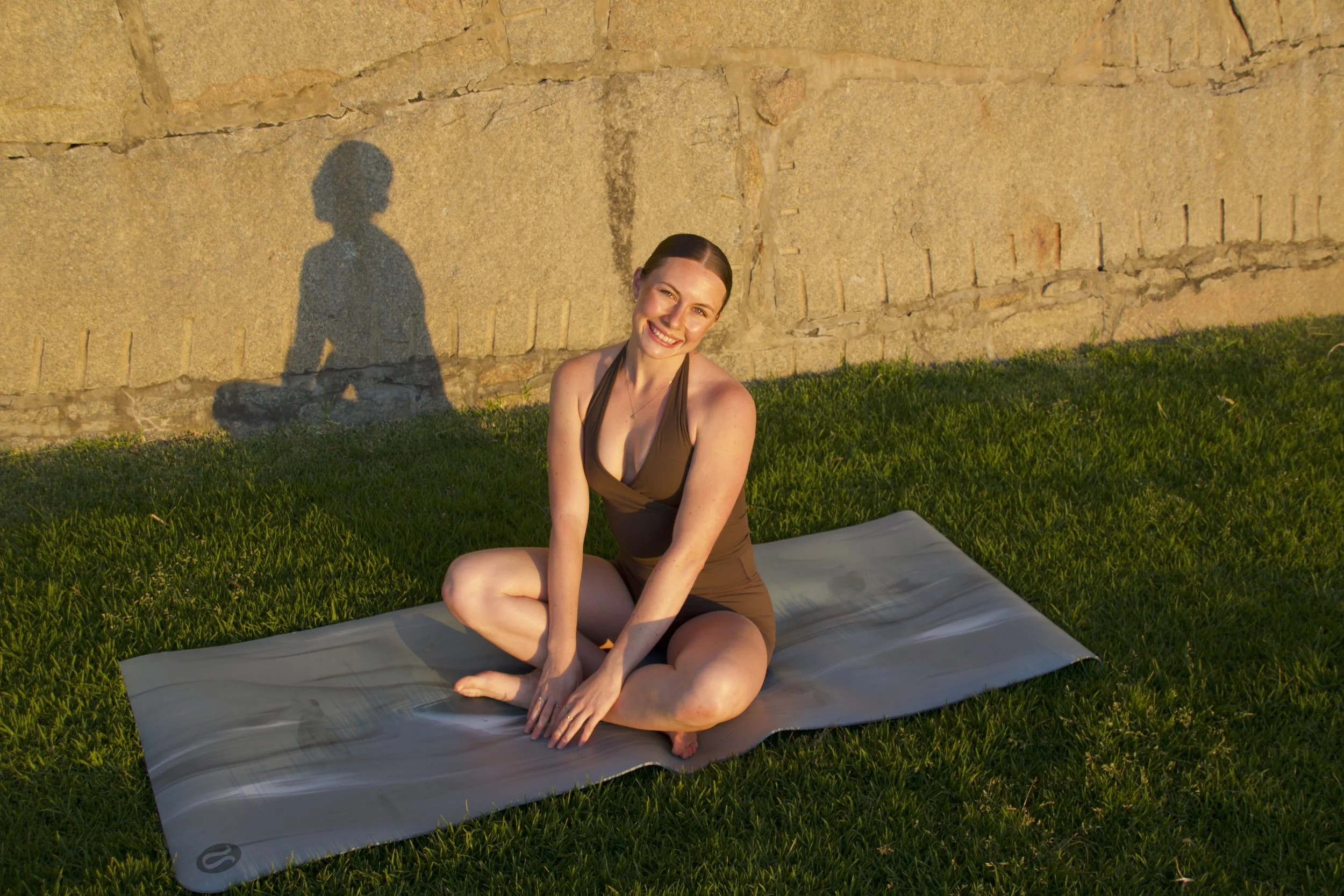 A woman sitting cross-legged on a yoga mat outdoors, smiling at sunset, with her shadow cast on a stone wall behind her.