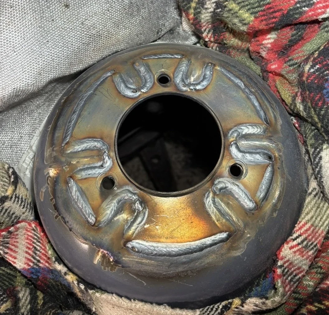 A metal wheel hub with multiple weld beads arranged evenly around the center hole, resting on a fabric surface.