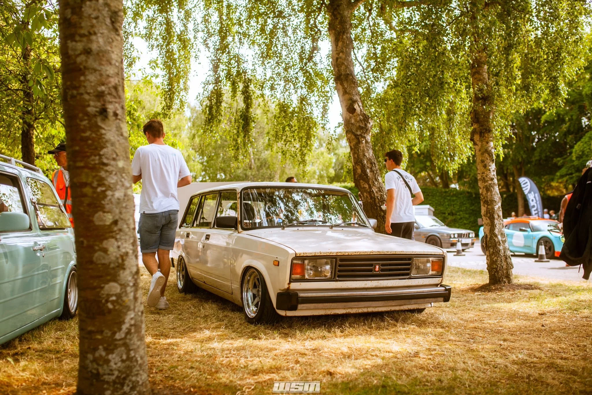 People walking around slammed lada riva estate at players classic under green trees at an outdoor event at goodwood motor circuit.