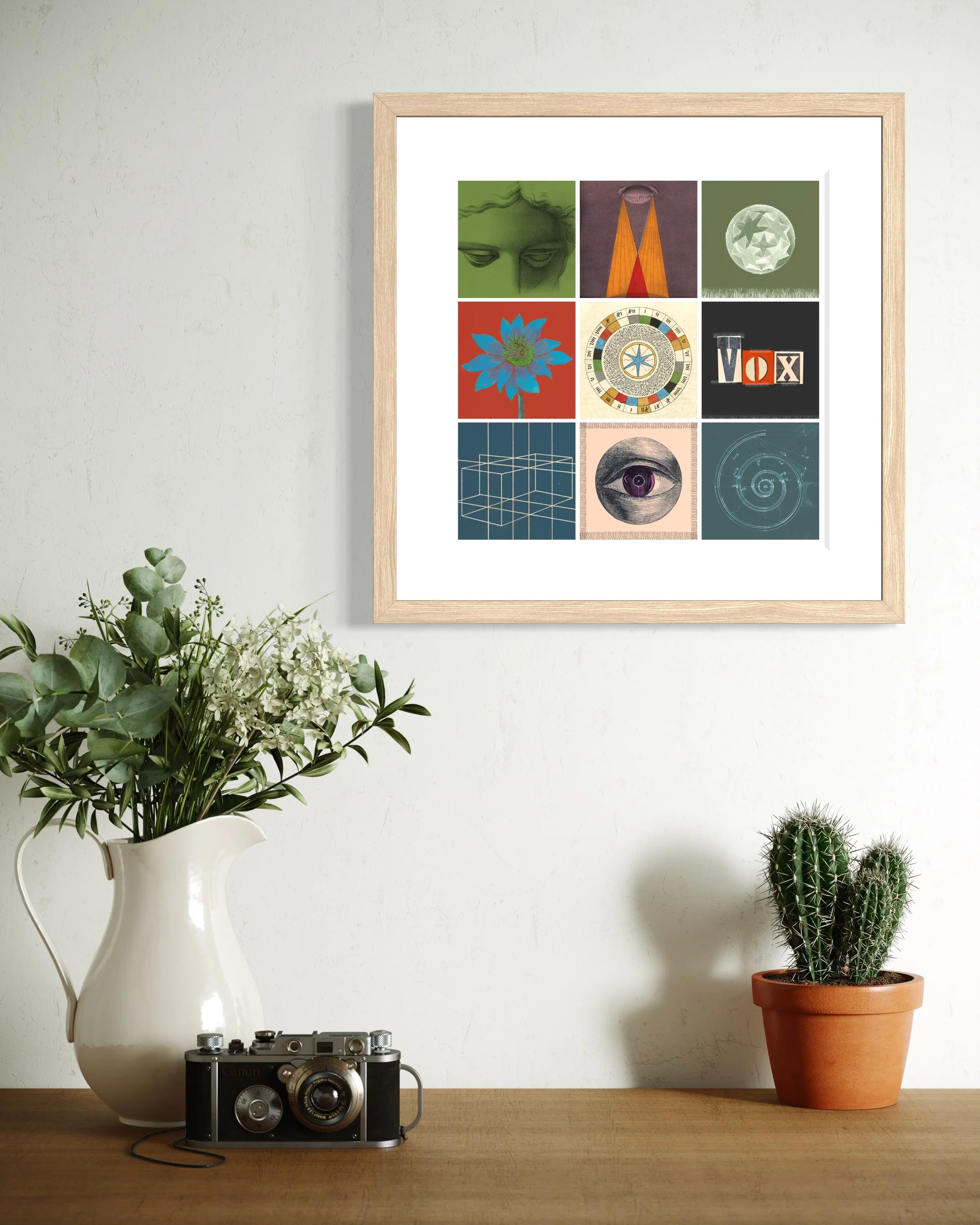 Alchemy fine-art print in an oak wood frame displayed on a wall above a wooden surface with a vintage camera, a white jug of flowers and a potted cactus
