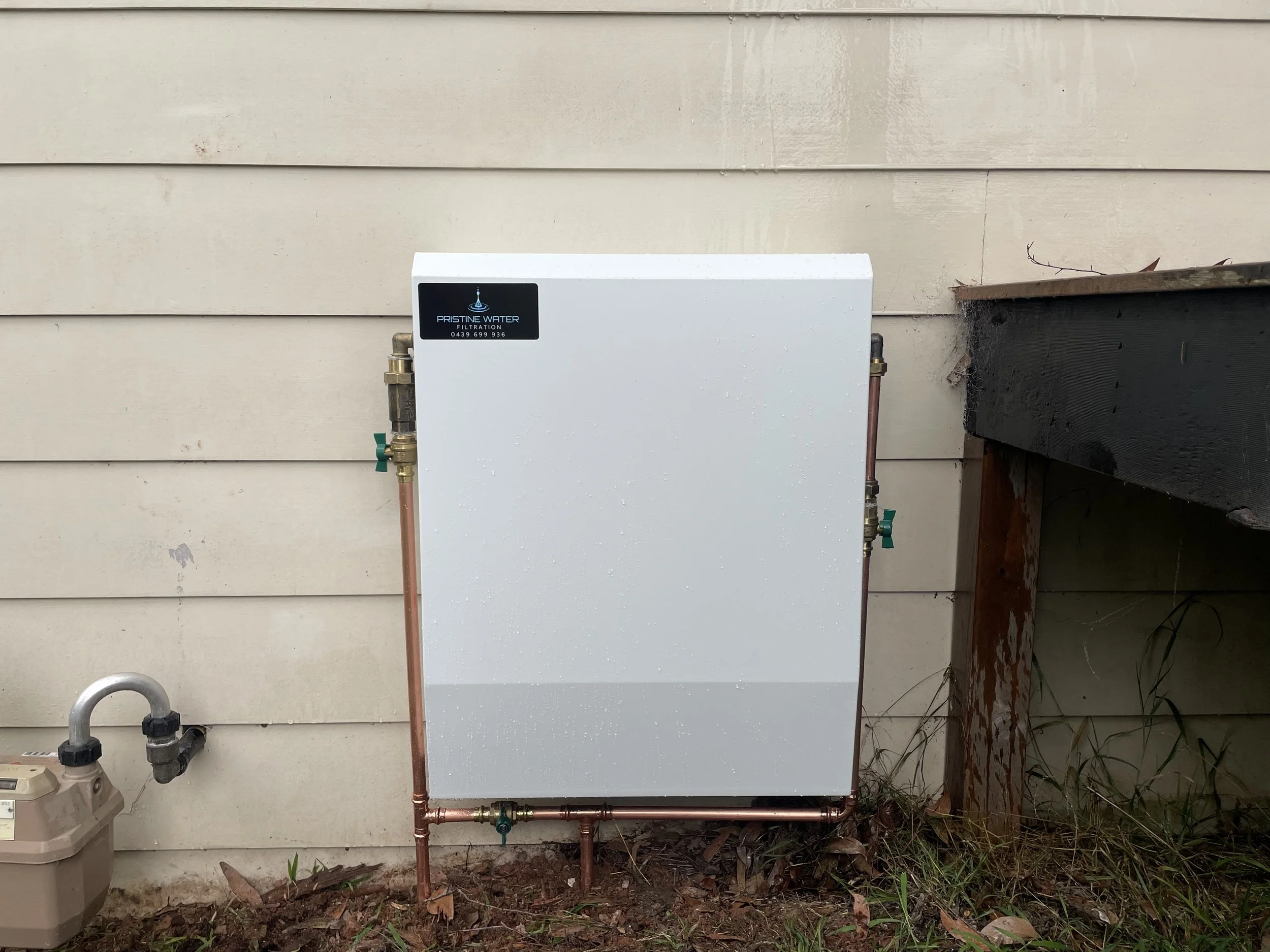 Outdoor water filtration system with copper pipes mounted on beige house siding, next to a dark wooden deck. There is a water meter on the ground to the left.