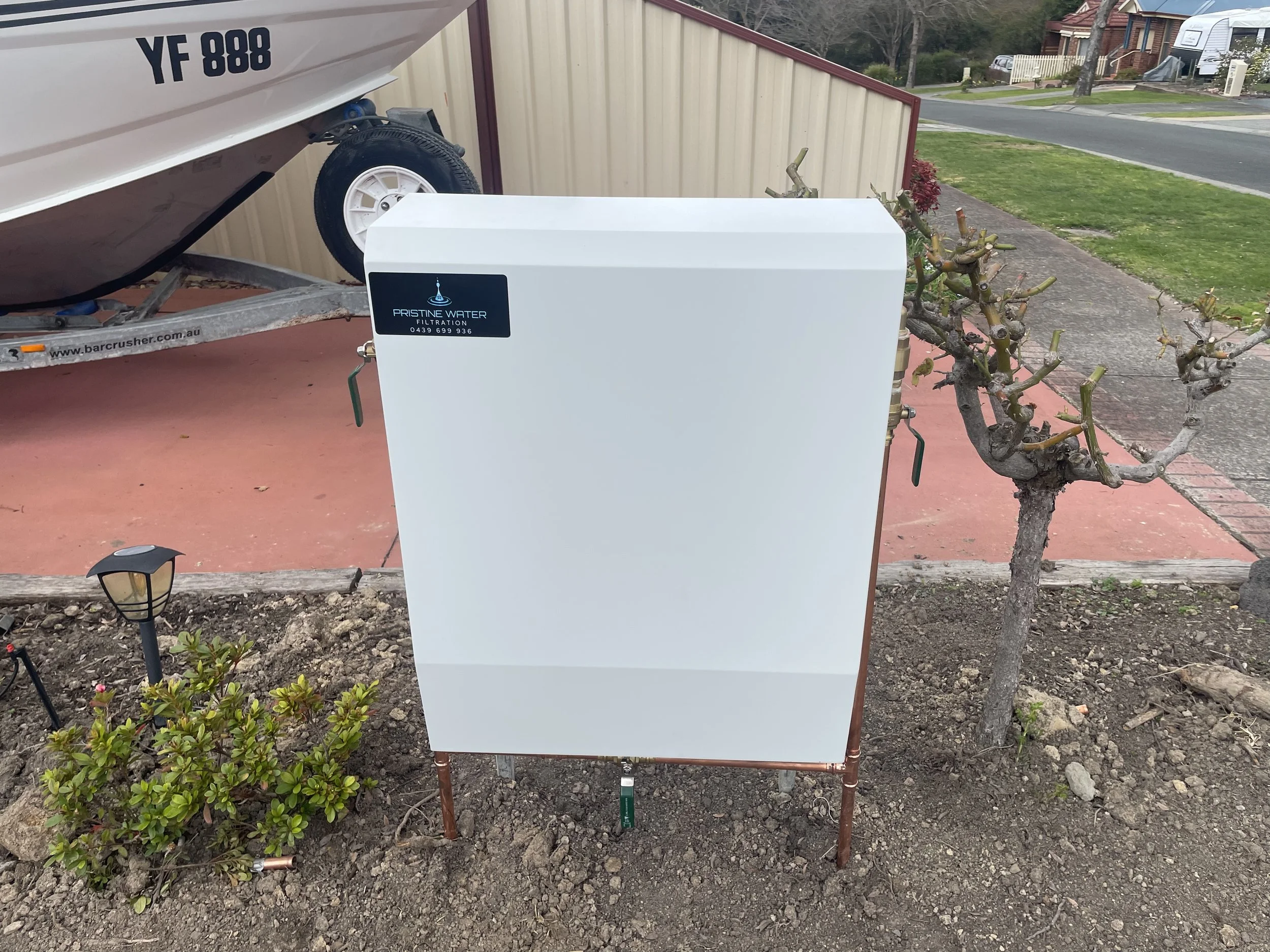 A white water filtration unit outdoors on a small patch of soil with a small tree, a bush, and a garden light in front. A boat on a trailer, a fence, and a street with houses are in the background.