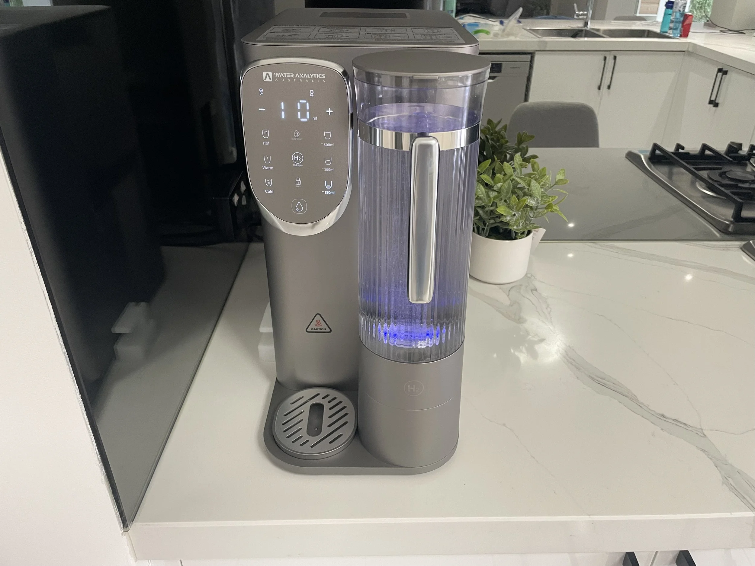 A water purifier with a digital display and glowing blue light on a kitchen countertop next to a potted plant, a stove, and kitchen cabinets.