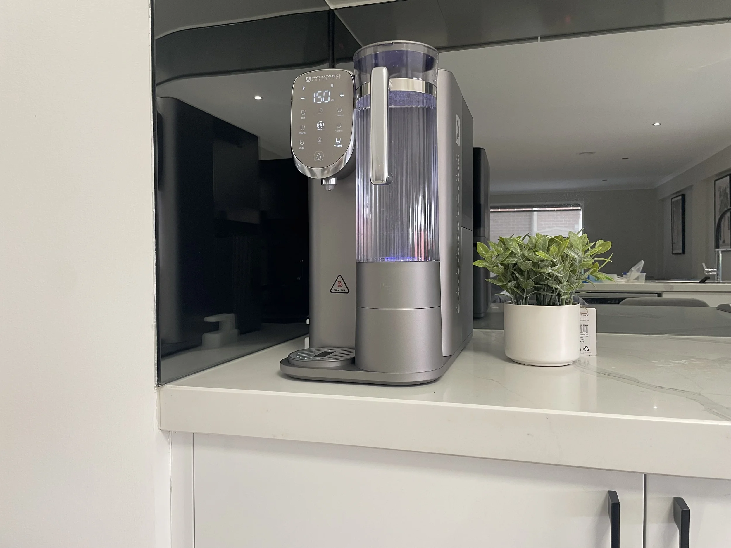 A modern water dispenser with a digital control panel displaying temperature, next to a small potted plant on a marble countertop in a bright kitchen.