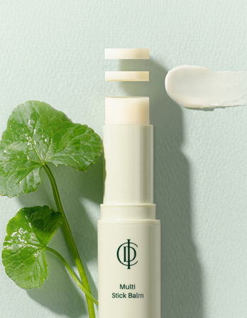 Tube of RIMAN multi stick balm with a white and cream colour palette, a green leaf to the left, and a smear of balm on the wall to the right.