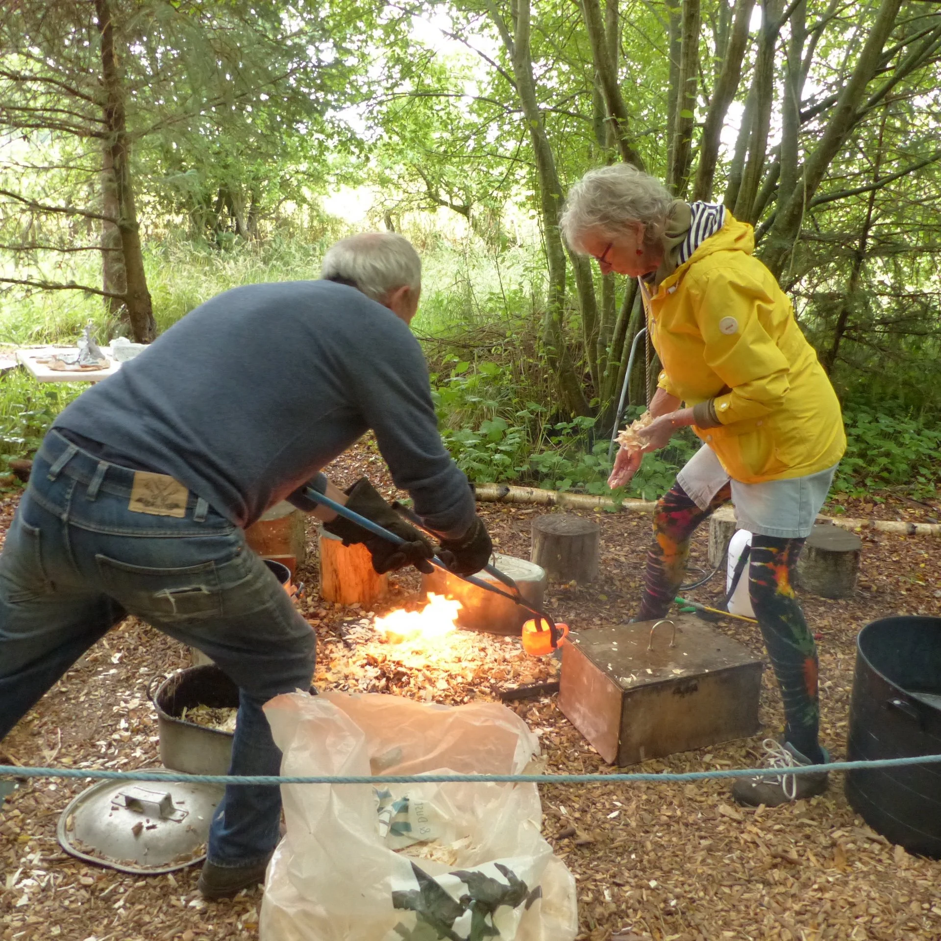 Two people working with molten glass outdoors in a wooded area, one is heating glass over a small fire while the other is shaping it using tools.
