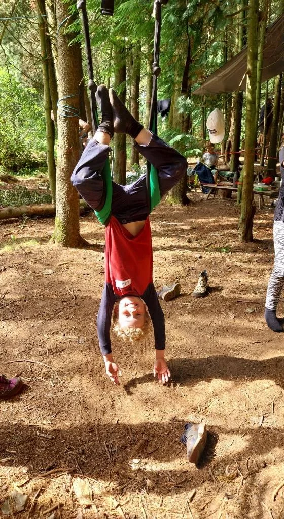 Child hanging upside down from a tree branch in a forest, holding a small ball while surrounded by trees and outdoor camping gear.