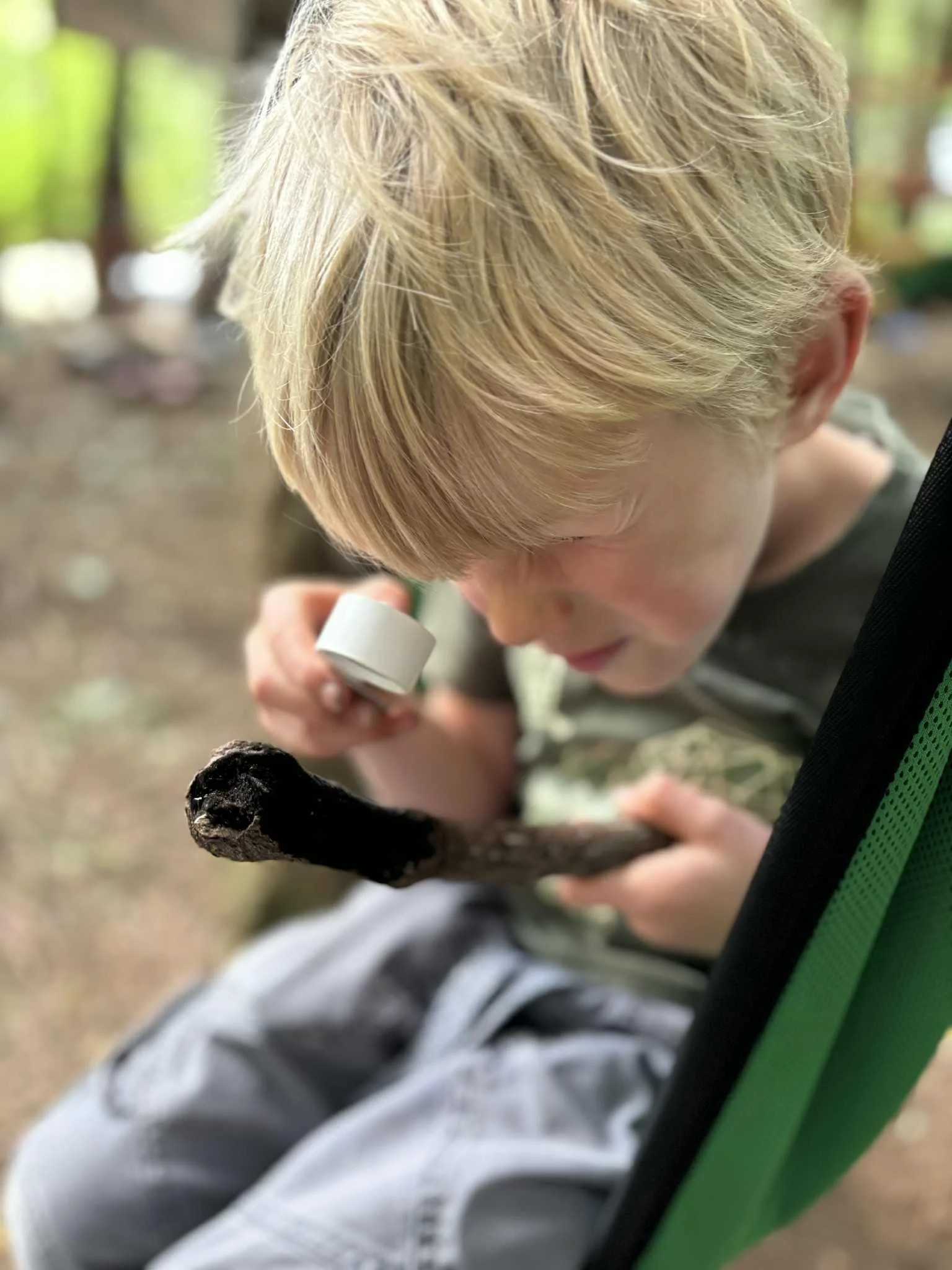 A young boy with blond hair sitting outdoors, closely inspecting a blackened stick or branch with a magnifying glass in his right hand. He appears to be examining or studying the object carefully.