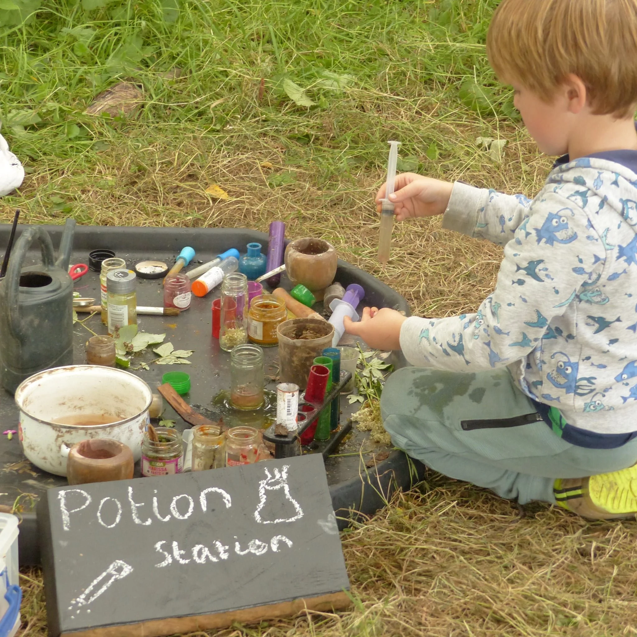 A young boy squatting outdoors on grass, conducting a science experiment with various jars and containers, with a sign reading 'Potion Station' in front.