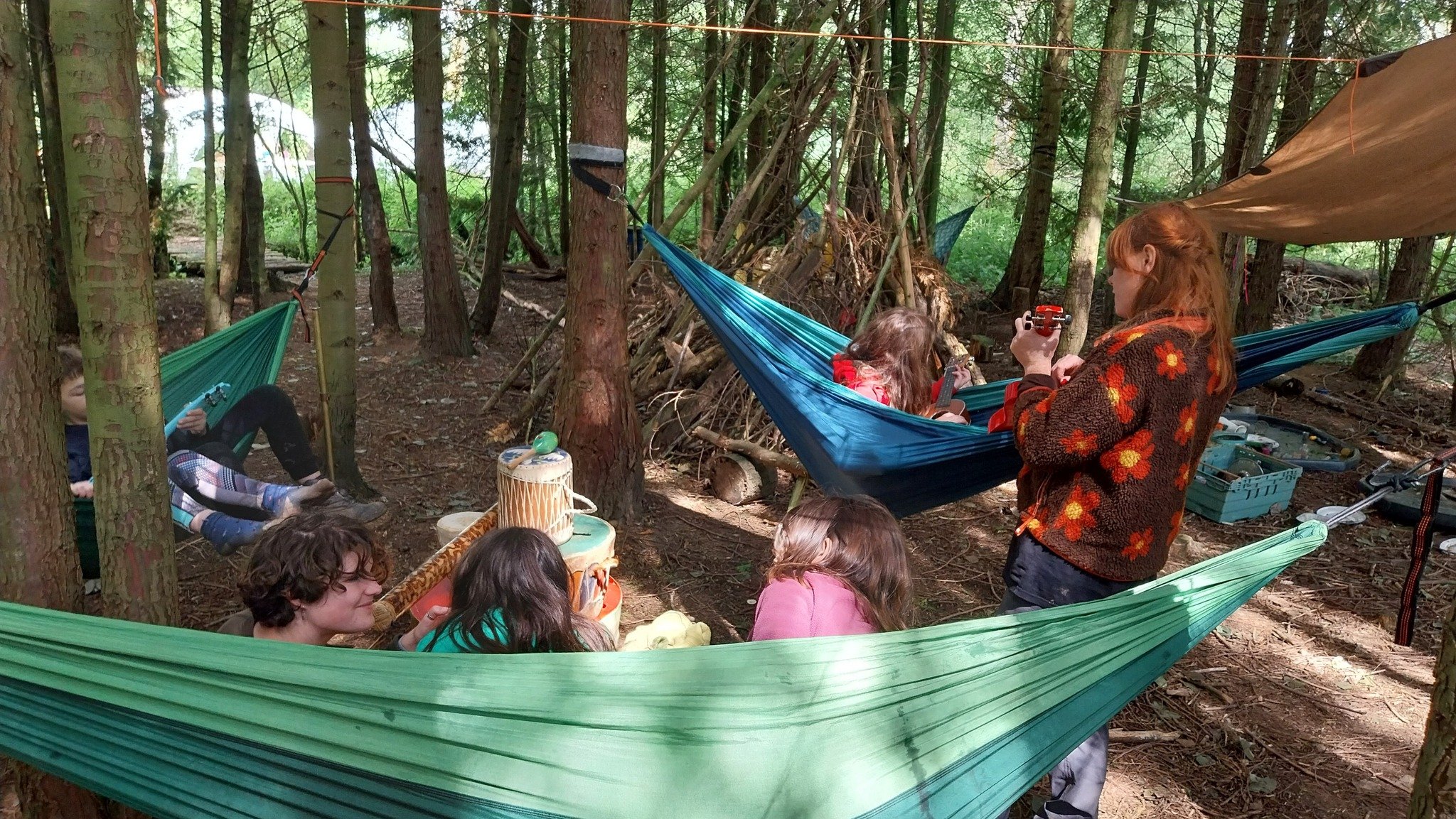 A group of children and a woman with red hair in a forest clearing, some lying in hammocks and others sitting or standing, enjoying a camping activity surrounded by trees and natural logs.