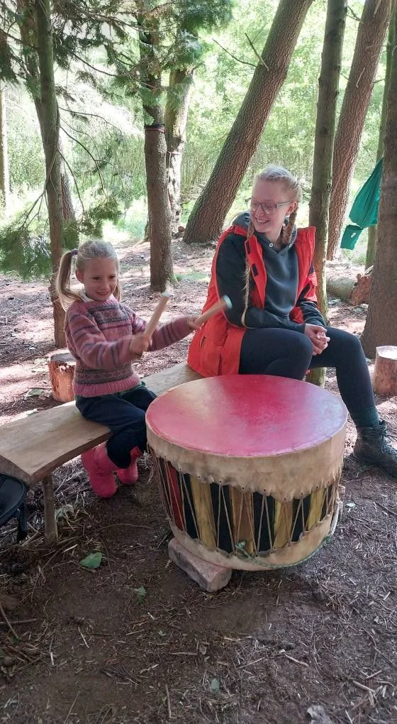 A young girl and an older girl sitting on a wooden bench in a forest, smiling and playing drums together. The girl on the left holds drumsticks, and the drum has a red top with a decorative body. All are seated outdoors among trees, with sunlight fil