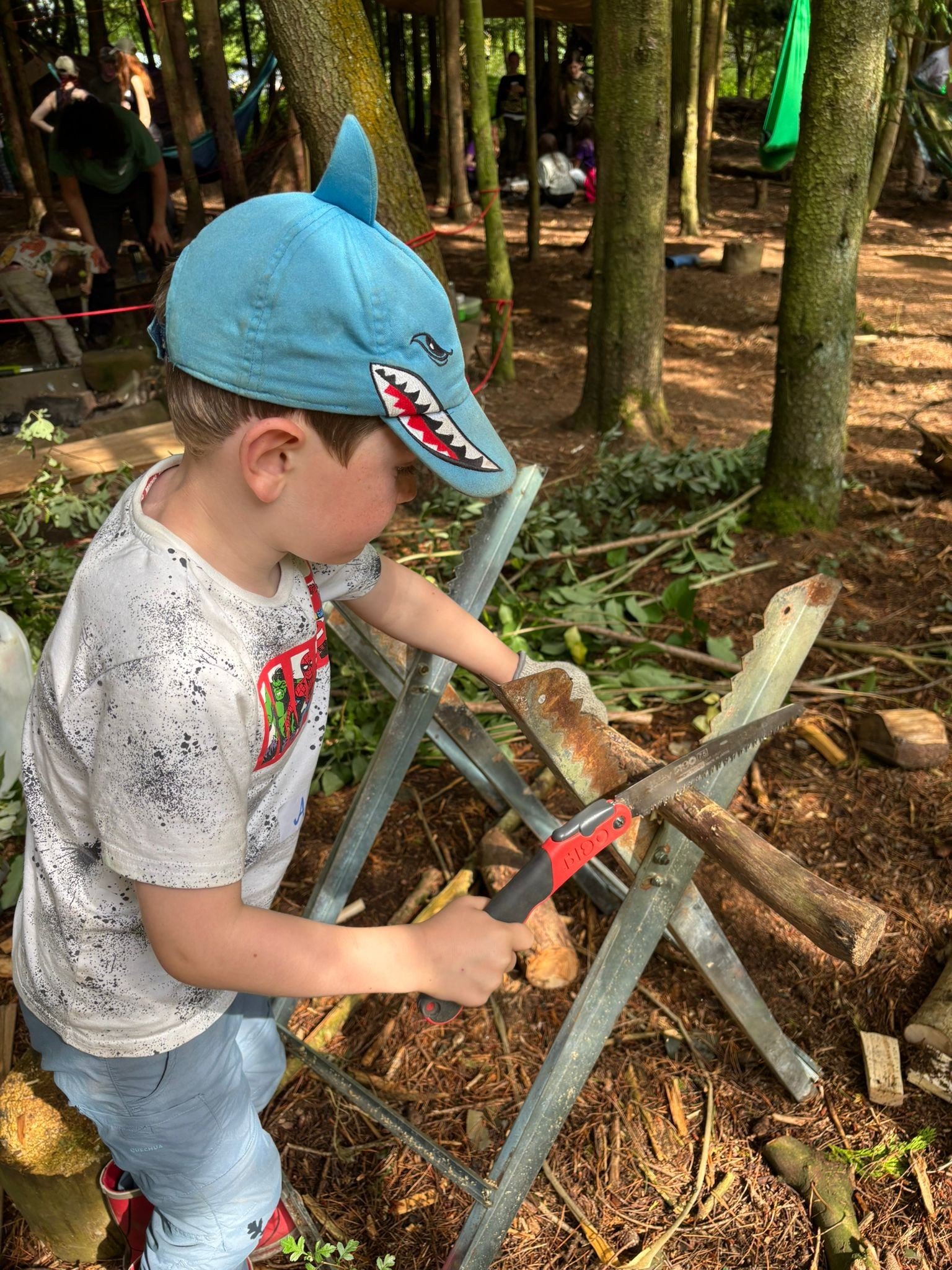 A young boy wearing a shark-themed blue cap and a white t-shirt is chopping wood with a saw in a forested outdoor setting.