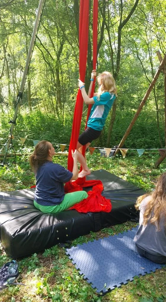 A girl with blonde hair wearing a blue and white shirt is climbing a red silk trapeze in an outdoor wooded area. An adult woman with brown hair, wearing a navy shirt and green pants, supports her by holding her legs. They are on a thick padded mat, w