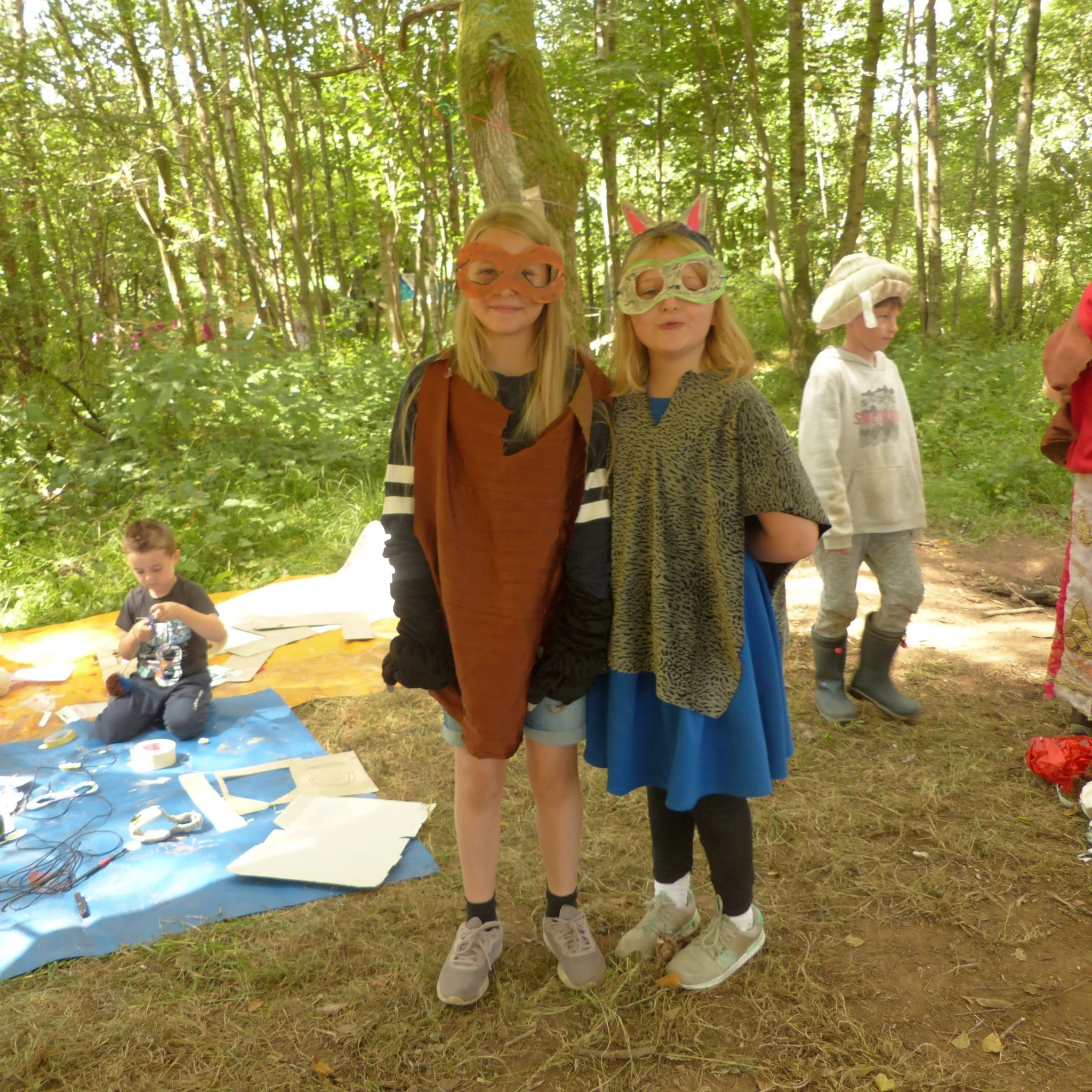 Two children dressed as animals with masks and costumes in a forest during daylight, with kids and craft supplies in the background.