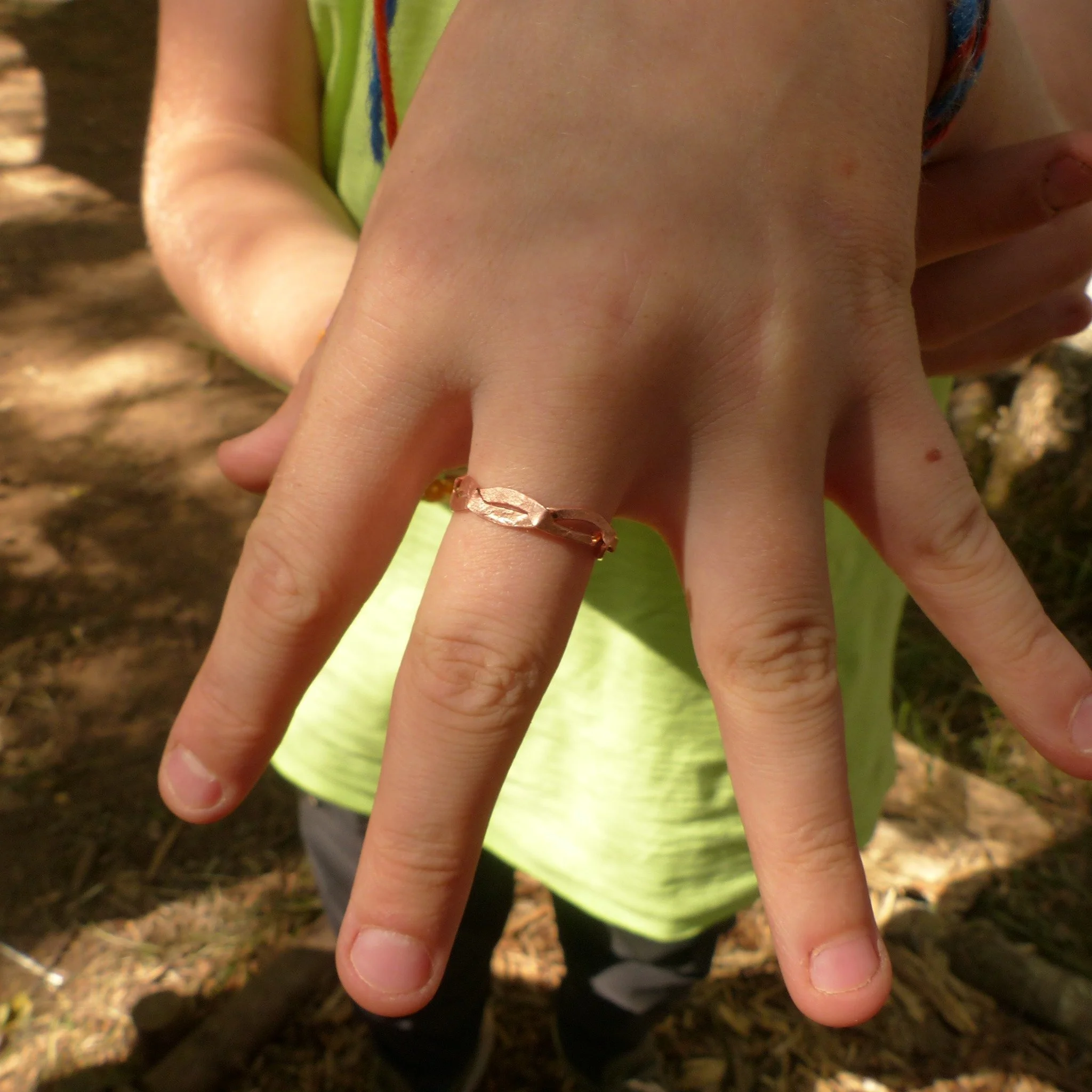 Person showing their left hand with fingers spread apart, wearing a simple copper ring on the middle finger, outdoors with trees and sunlight in background.
