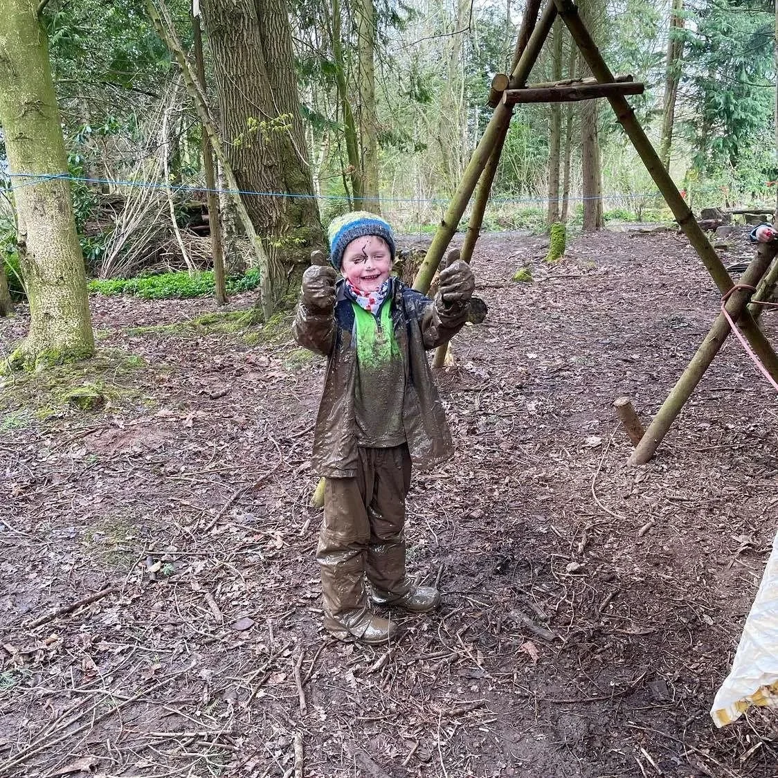 A young boy in muddy clothes, smiling, standing in a muddy forest clearing during daytime, in front of a wooden A-frame structure.