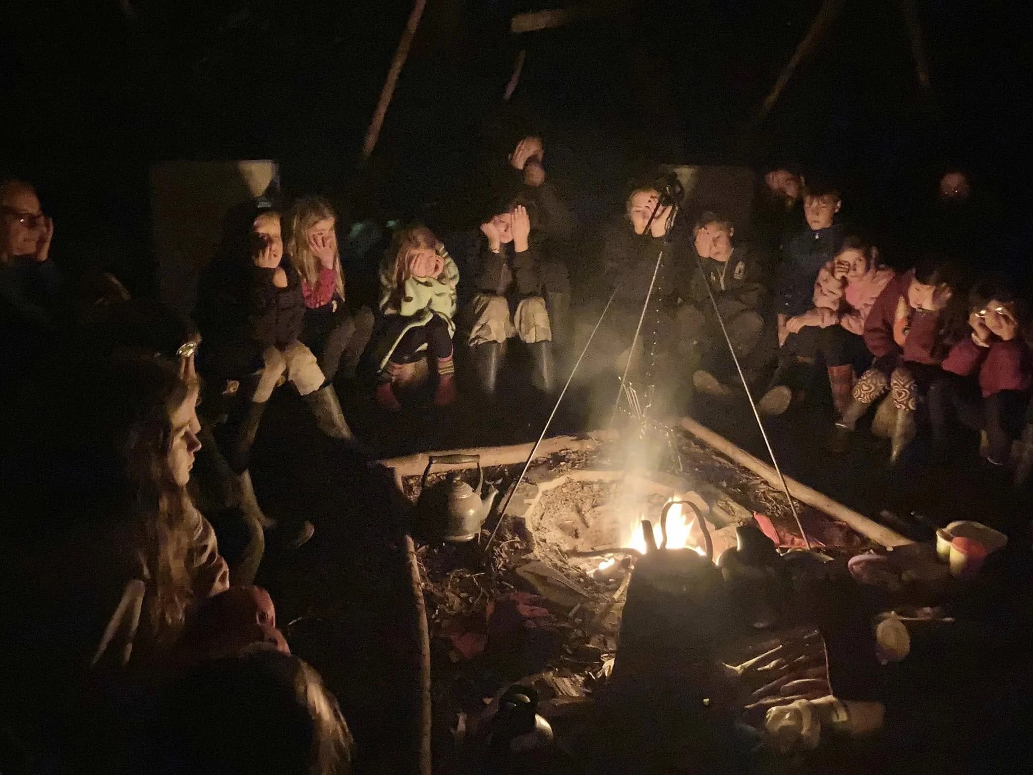 A group of children and adults sitting around a campfire inside a dark tent, appearing to be listening or watching something with expressions of amazement or surprise.