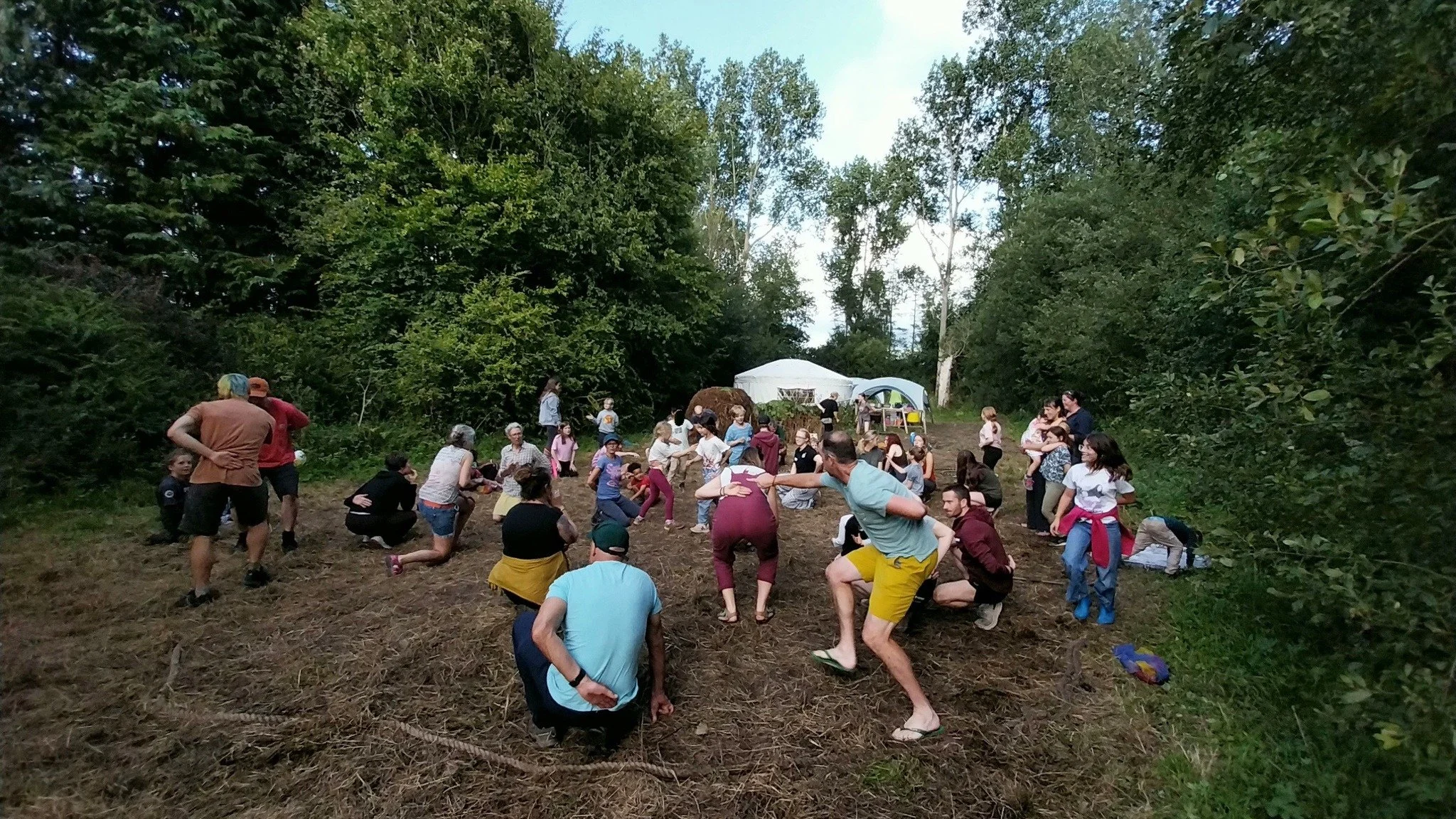 People gathering and playing in an outdoor wooded area during daytime, with tents and a hay bale in the background.