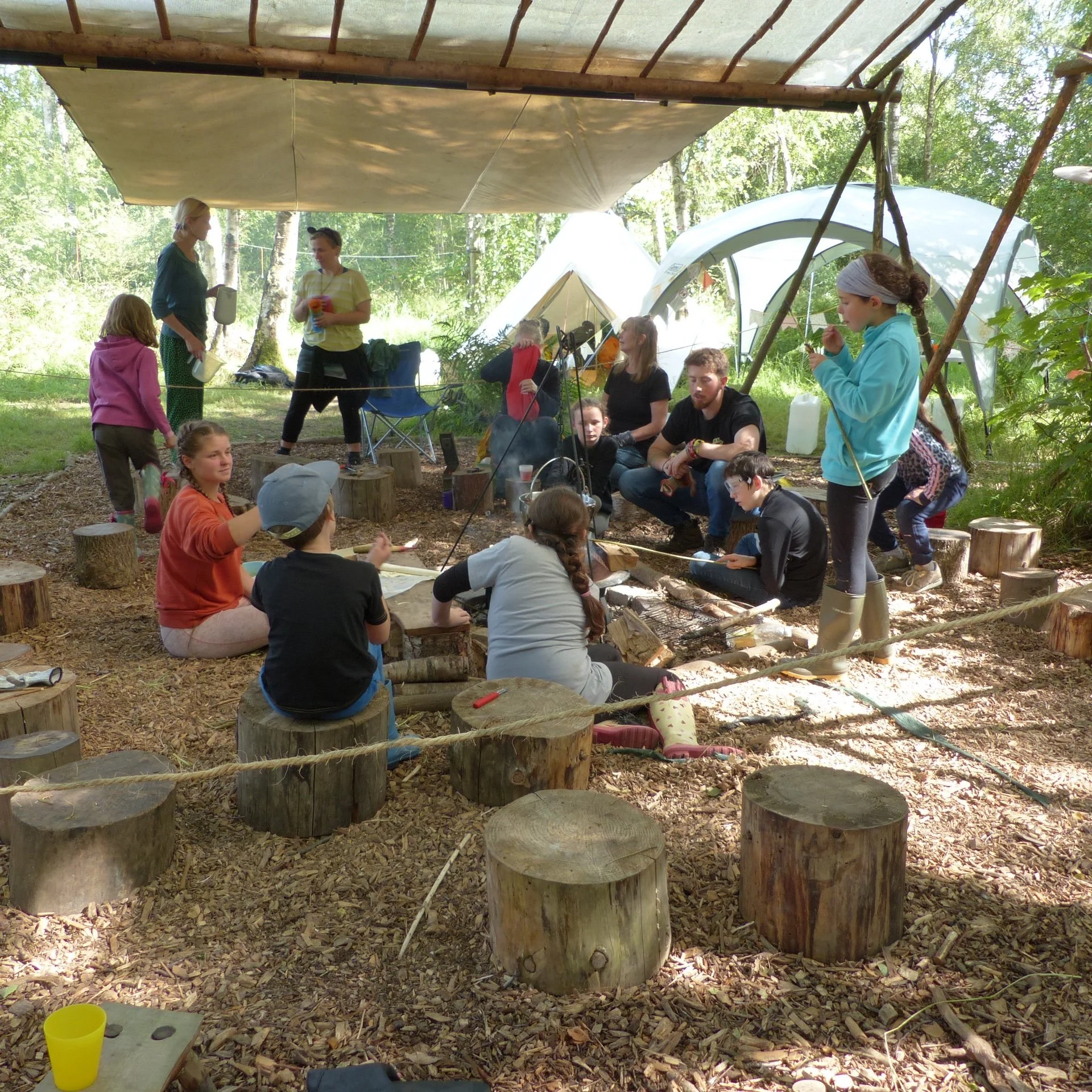 Group of children and adults gathered around a campfire in a wooded outdoor setting for a campfire program.