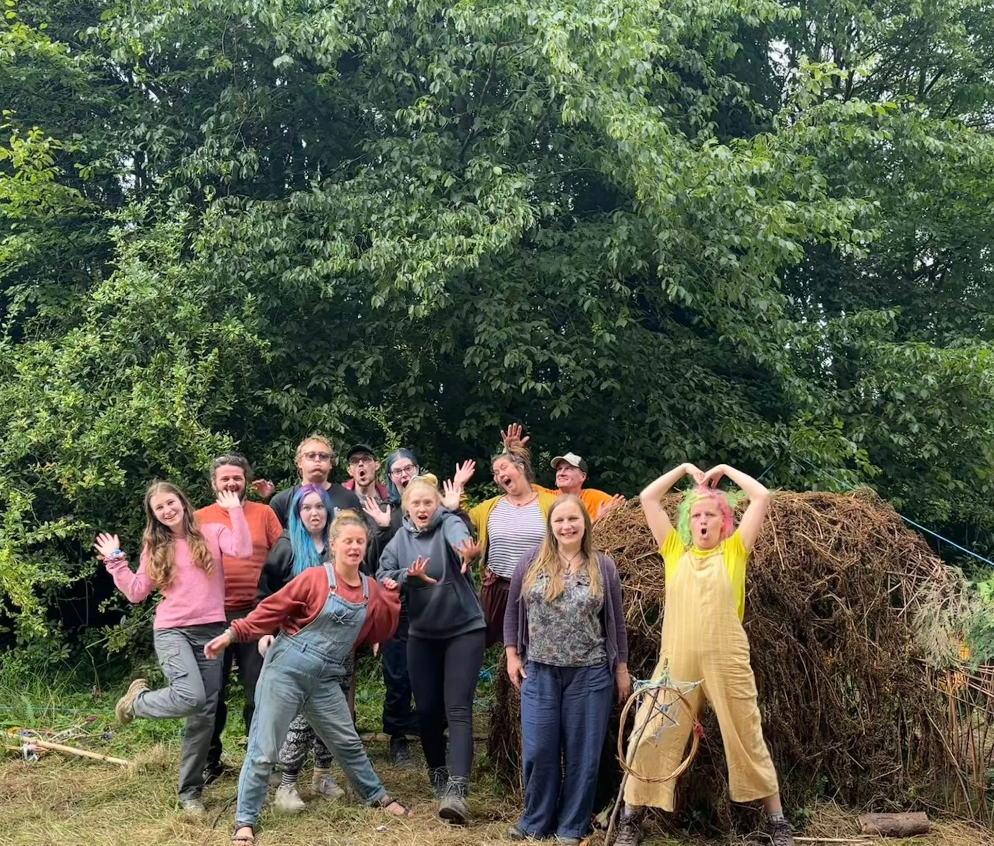 Group of people posing outdoors in front of a large pile of branches and foliage, with trees in the background, some making playful or expressive gestures.