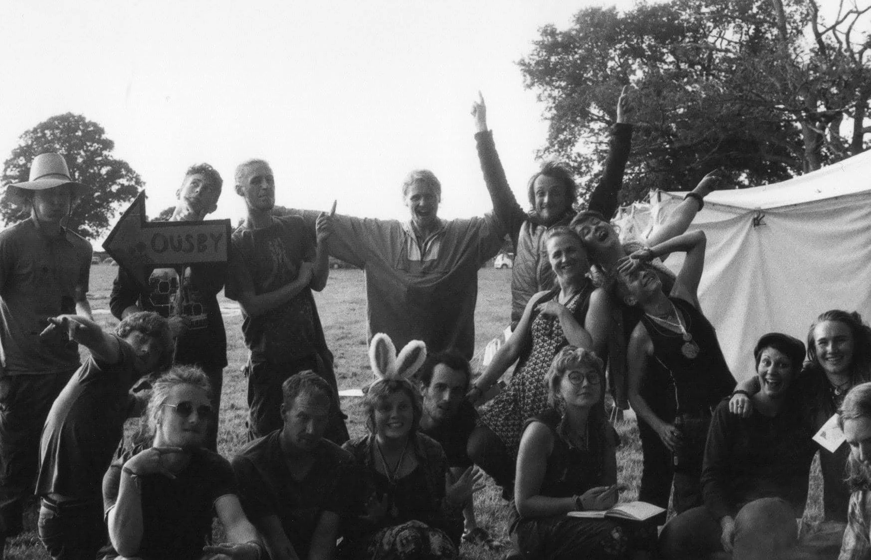 Group of people outdoors, some wearing costume accessories like bunny ears and medals, posing for a photo during an event or festival, with trees and tents in the background.