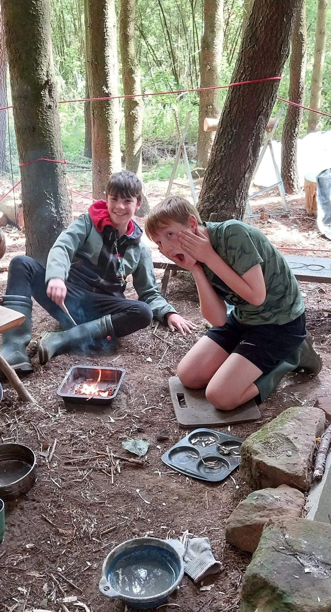 Two young boys are camping in the woods. One boy is kneeling on a cutting board and covering his face, while the other boy is sitting on the ground smiling. There is a small fire in a metal tray, various camping supplies, and trees surrounding them.
