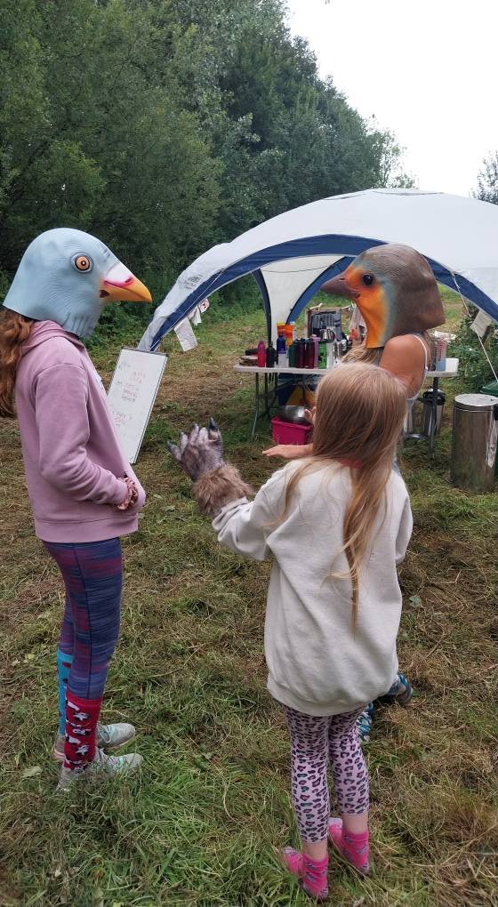 Two children in animal masks, one with a bird mask and the other with a duck mask, are talking outdoors near a tent, with a girl in a fleece jacket and leopard print leggings holding a raccoon.