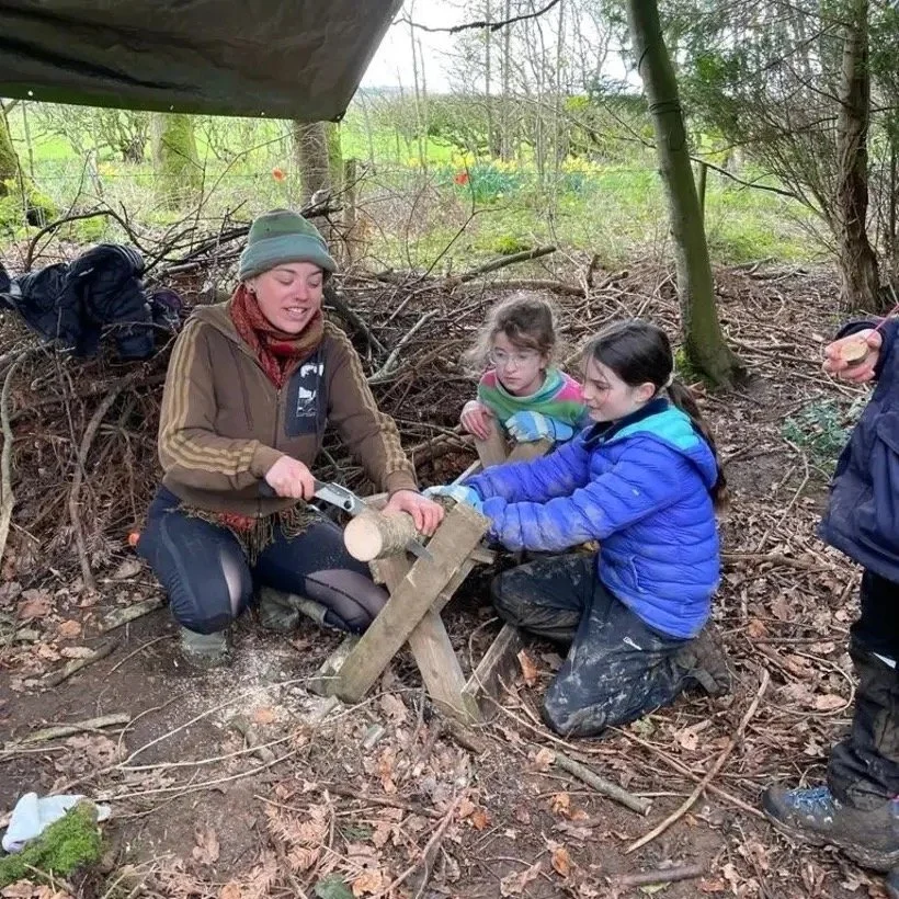 Three children and an adult woman in a forest, using a saw to cut a small log placed on a wooden workbench.