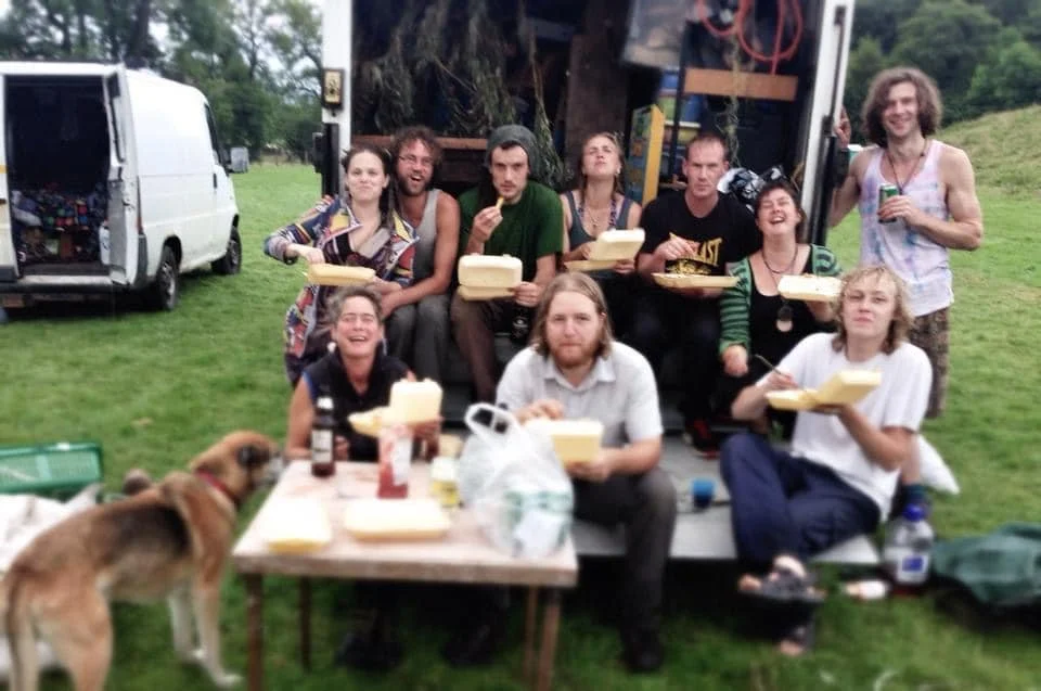 Group of people enjoying a picnic outdoors with pizza, drinks, and a dog in a grassy area near a white van and a truck.