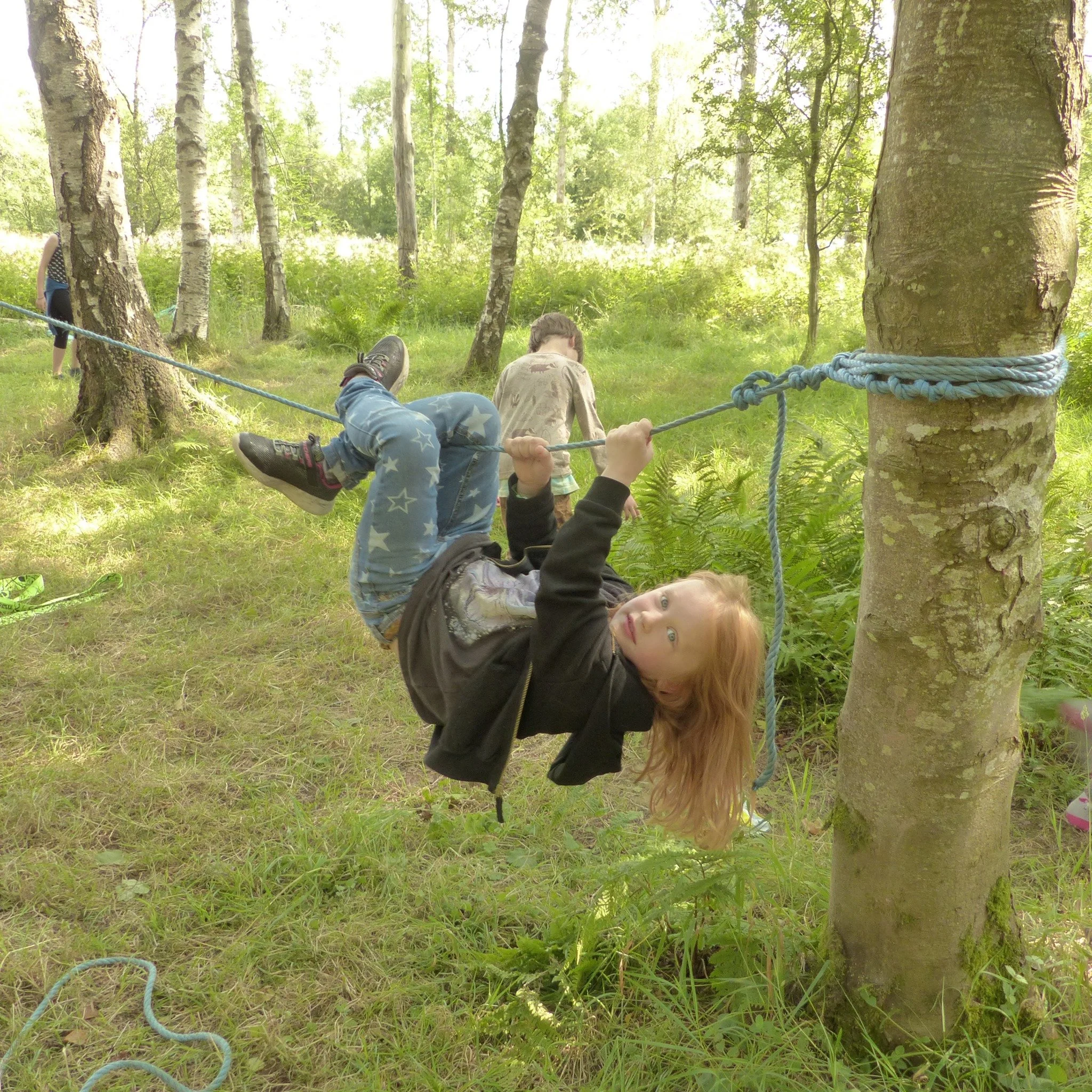 A young girl with long red hair and green eyes is hanging upside down on a rope tied between two trees in a forest, smiling and looking at the camera.