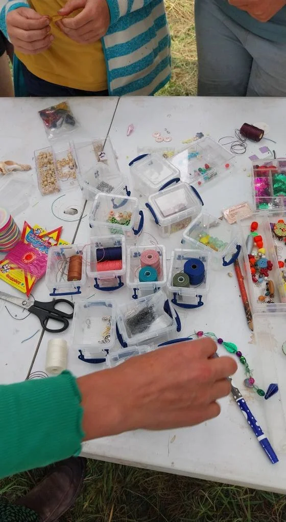 A white table with various craft supplies including spools of thread, beads, scissors, and small containers, with hands of people working on a jewelry project.