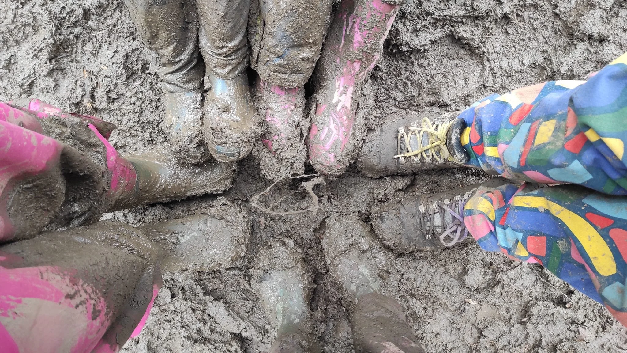 View of muddy ground with four people standing close together, their legs and feet visible. The ground is wet and covered in thick mud, with some parts caked on the shoes and pants. The people are wearing colorful pants and sneakers, which are also muddy.