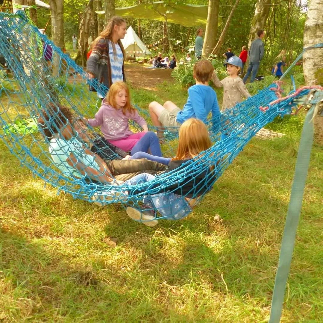 Children sitting and relaxing on a large blue hammock in a wooded outdoor area with adults and a tent in the background.