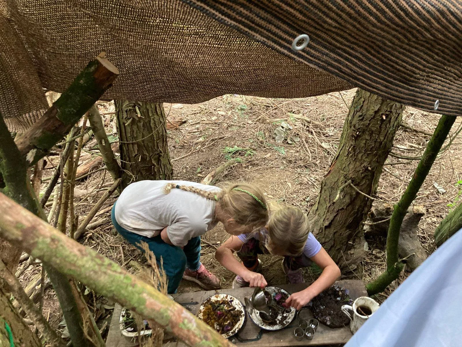 Two young girls with blonde hair, wearing casual clothes, are kneeling and leaning over a small wooden table outdoors in a forest, preparing breakfast or a meal with plates, cups, and utensils. They are surrounded by trees and branches.