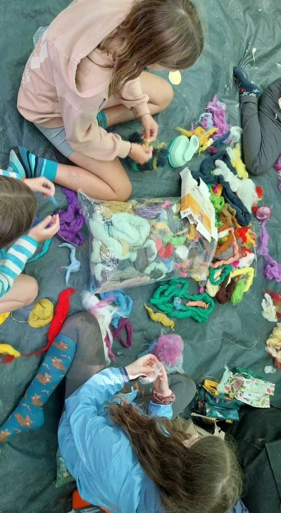 Children sitting on the floor, crafting with colorful yarn and wool, with a large plastic bag filled with more yarn and wool around them.