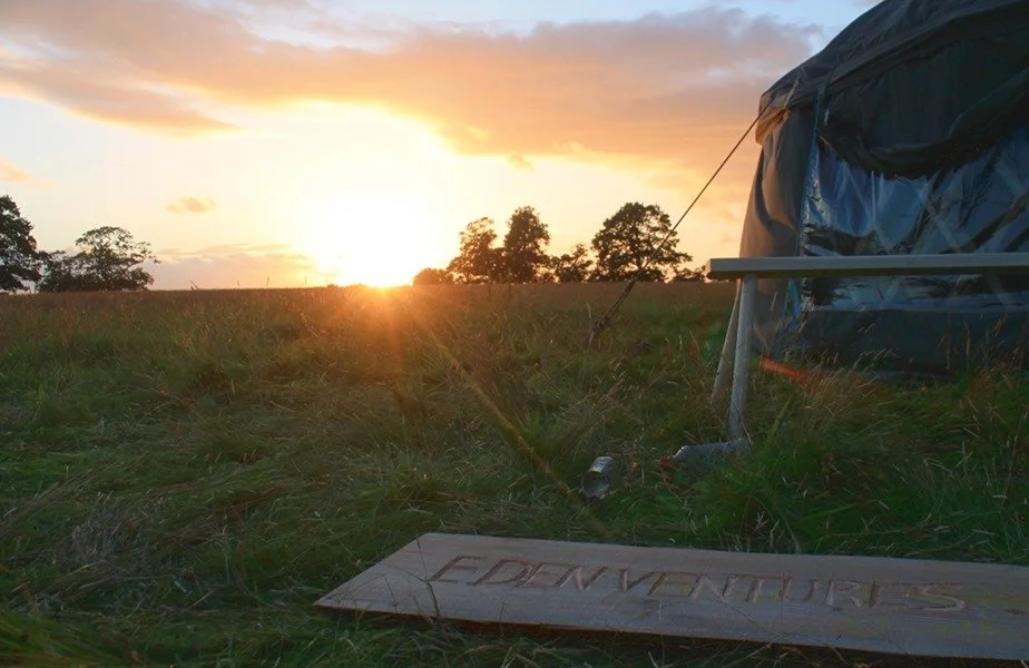 Sunset over a grassy field with trees in the distance, a tent on the right, and a wooden sign with the word "ADVENTURES" lying on the ground.