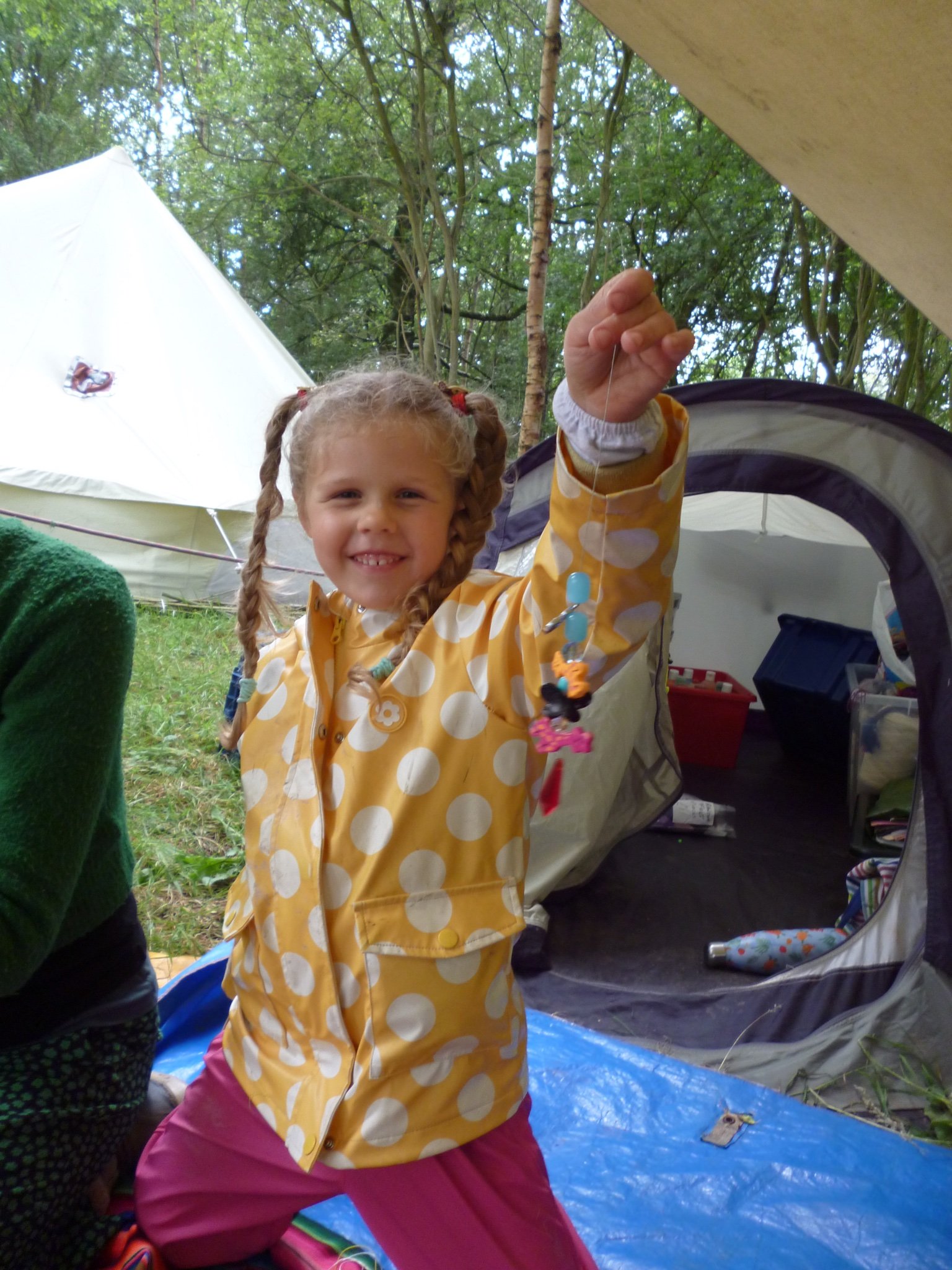 Young girl with curly hair in pigtails smiling and showing a string of colorful beads she made while camping outdoors. Tent and trees are visible in the background.