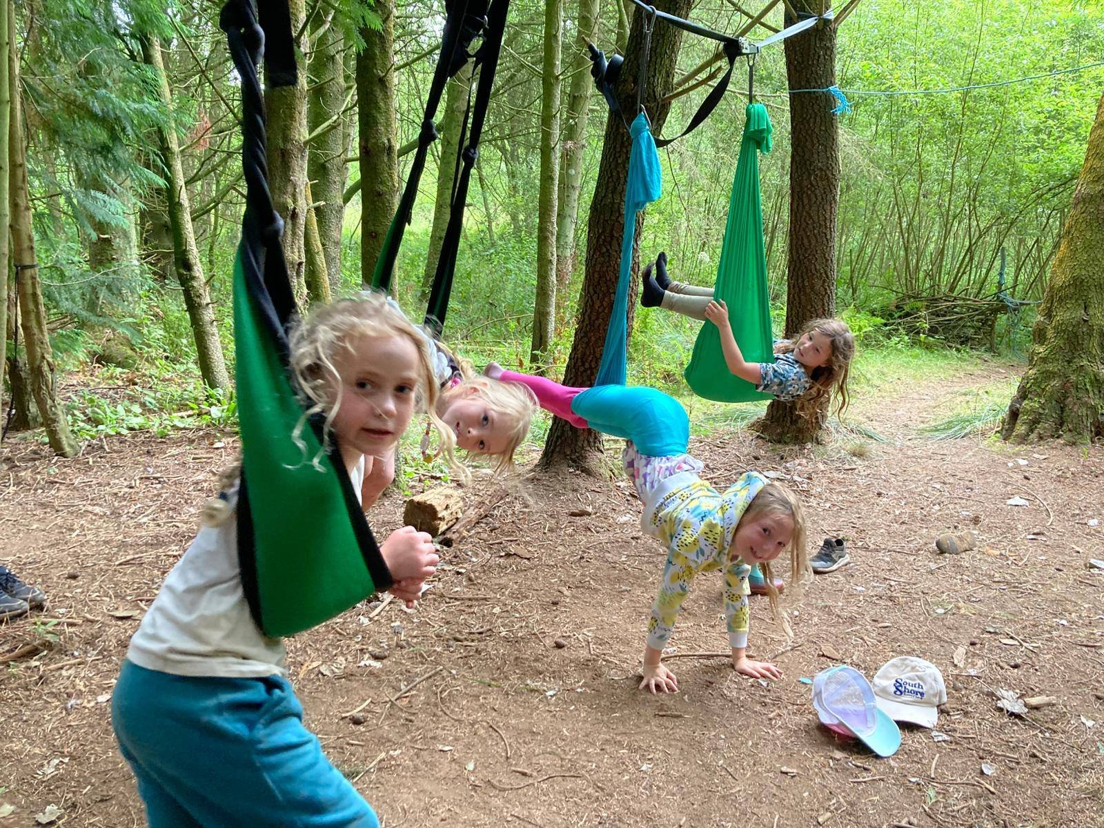 Four young girls playing on aerial silk swings in a wooded forest area. The girls are hanging upside down and on their hands and knees on the ground, with trees and forest greenery in the background. There are caps and shoes on the ground nearby.