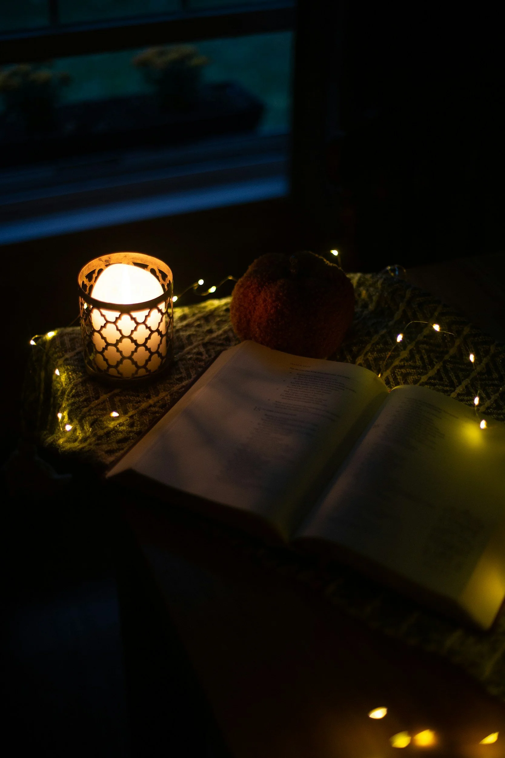 Open Bible on a table illuminated by a candle and string lights, with a decorative fruit and a patterned cloth, in a dimly lit room.