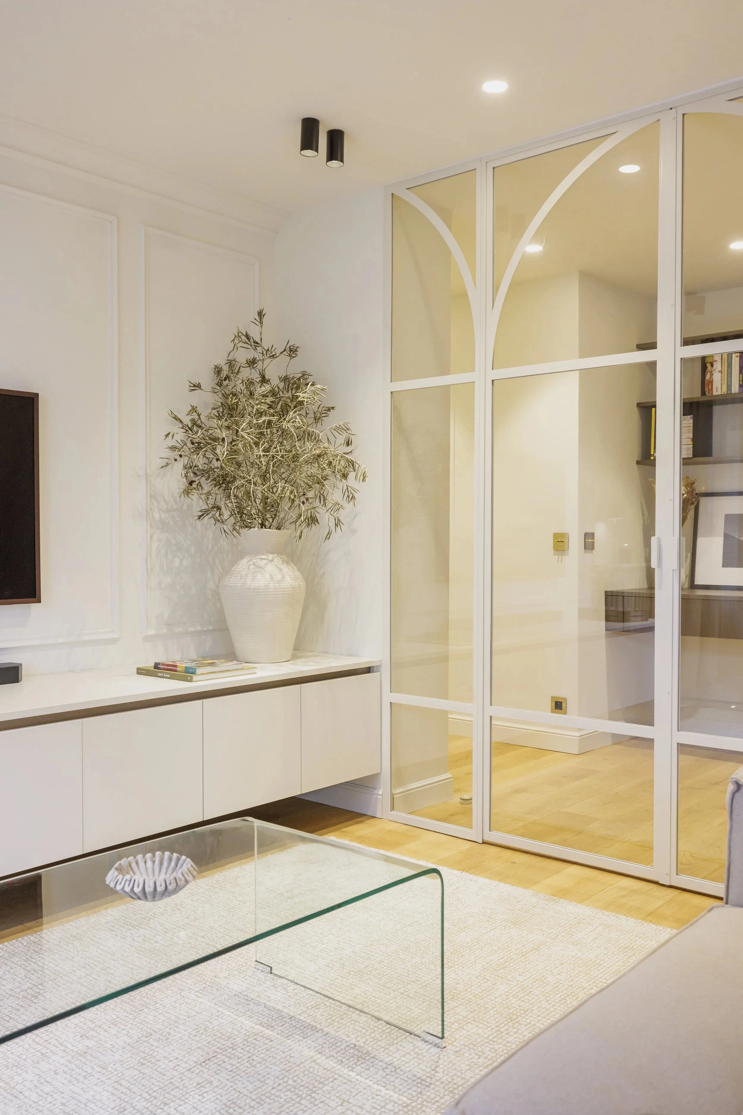 Salon moderne avec un mur blanc, une grande plante dans un vase blanc, un meuble blanc, une table en verre, un tapis beige, et une séparation vitrée blanc.