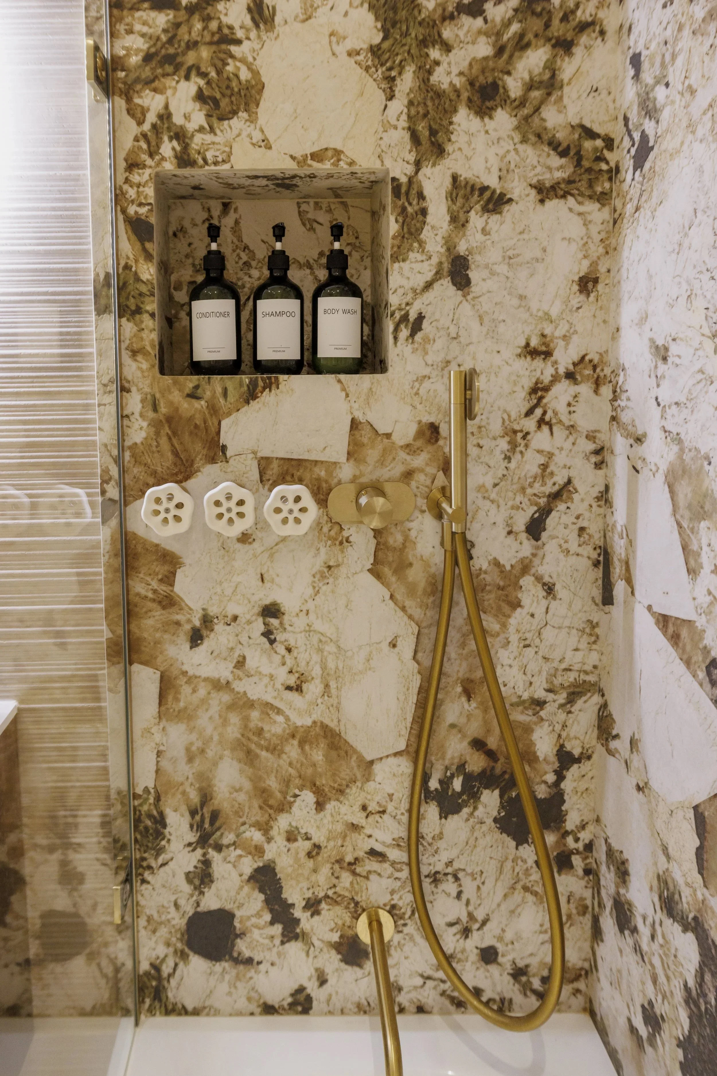 Une douche avec des bouteilles de produits de toilette (conditionneur, shampoing, gel douche) posées dans un nichoir intégré dans le mur en marbre beige et marron.
