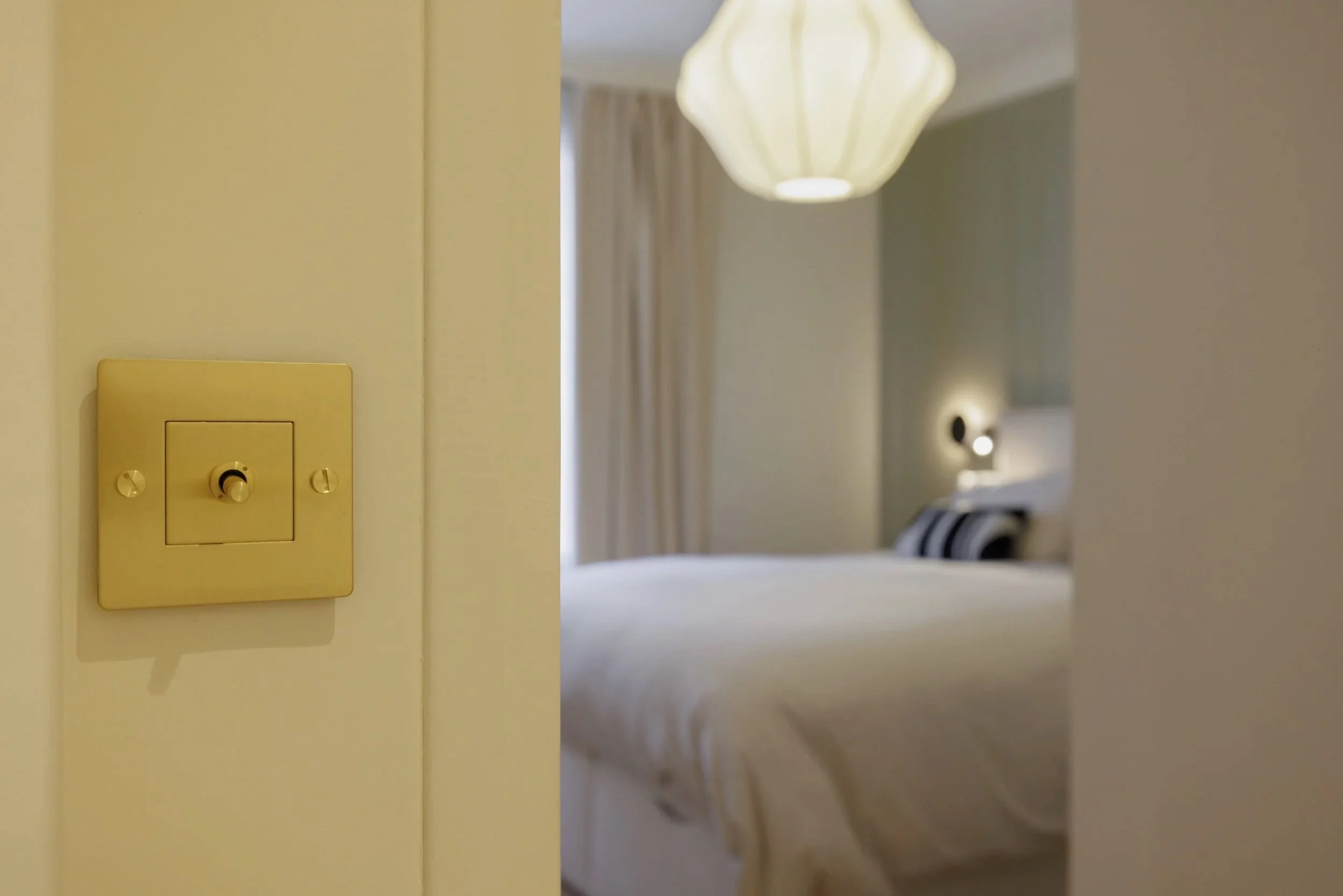 Vue partielle d'une chambre d'hôtel avec un lit blanc, une lampe suspendue au plafond et une lumière de lecture allumée, vue à travers une porte entrouverte.