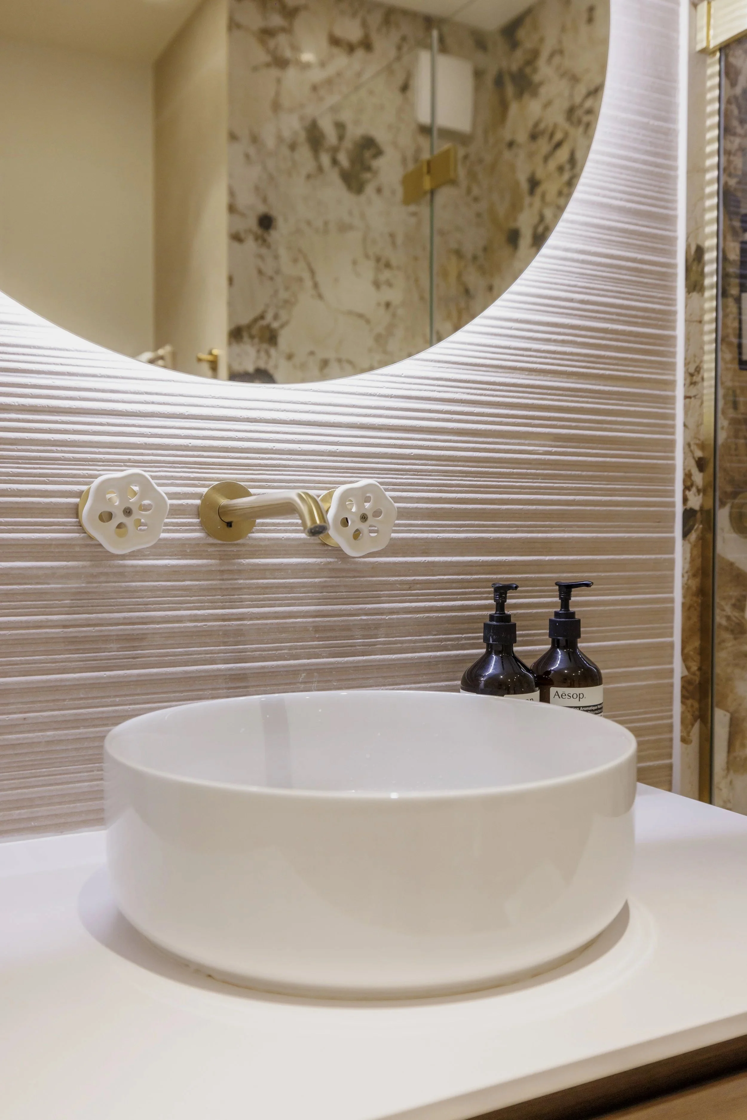 Un lavabo blanc rond en porcelaine avec deux bouteilles de savon Aesop sur la droite, un miroir rond au-dessus, une robinetterie ancienne en laiton avec poignées en forme de fleurs, murs en pierre et carreaux muraux texturés en beige.