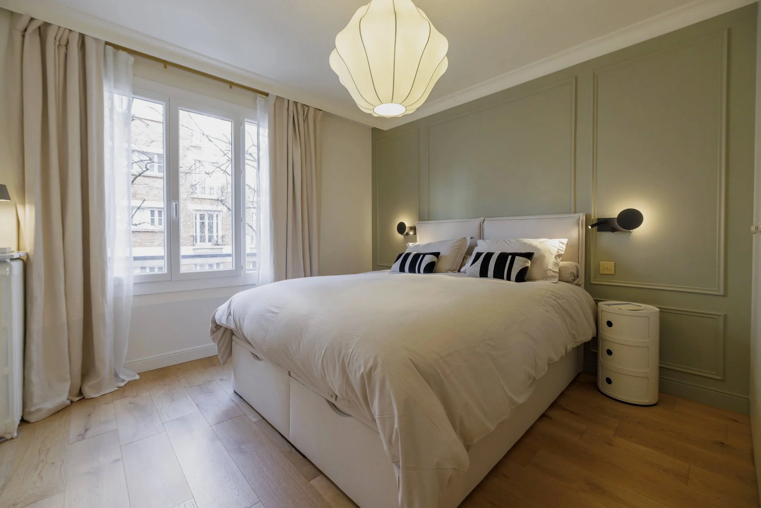Chambre avec lit double, draps blancs, coussins noirs et blanc, lampe suspendue au plafond, fenêtre avec rideaux blancs, vue d'arbres et bâtiments